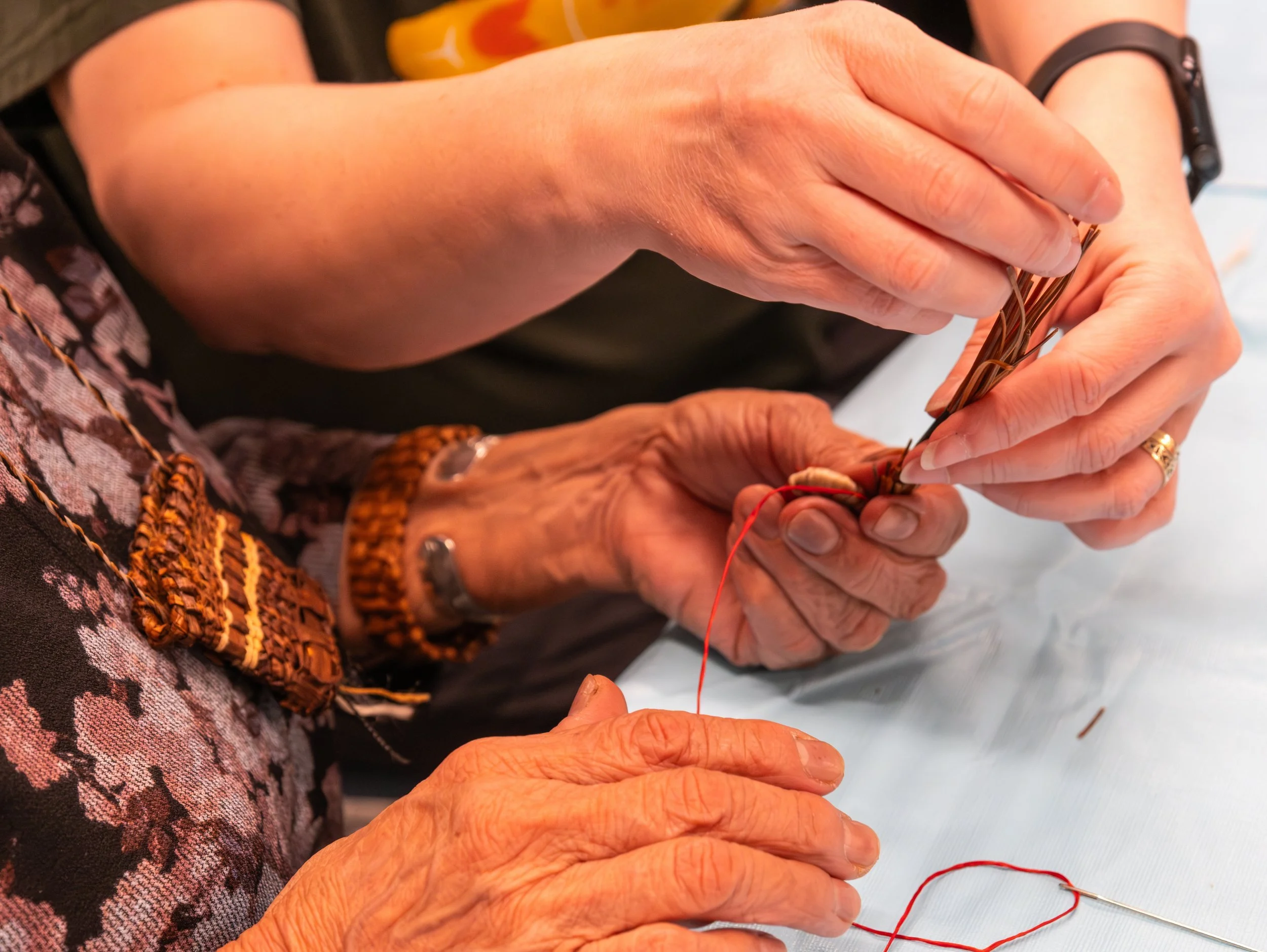 Close-up of two sets of hands sewing red thread around a bunch of pine needles.