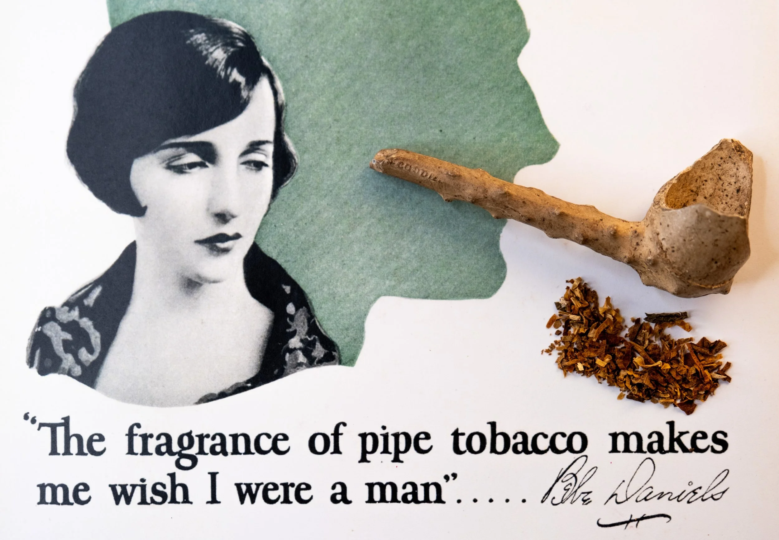 A vintage illustration of a woman with dark hair, alongside a green silhouette of a face in profile. There is a pipe, loose tobacco, and a quote that says, "The fragrance of pipe tobacco makes me wish I were a man" attributed to Bebe Daniels.