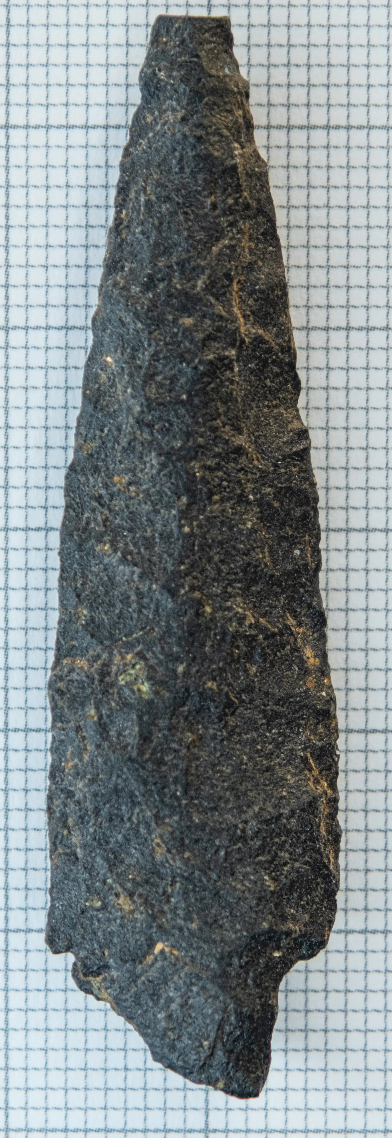 Close-up of a dark, triangular stone tool with a pointed tip, placed on a graph paper background for scale. This is an ancestral Coast Salish spear point.