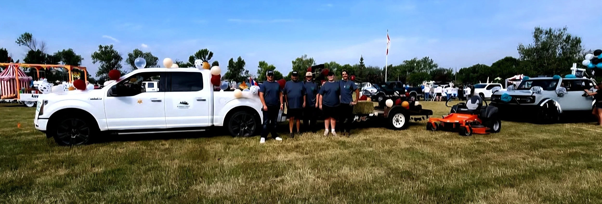 People standing in front of a decorated white pickup truck at an outdoor event, with other vehicles and tents in the background.