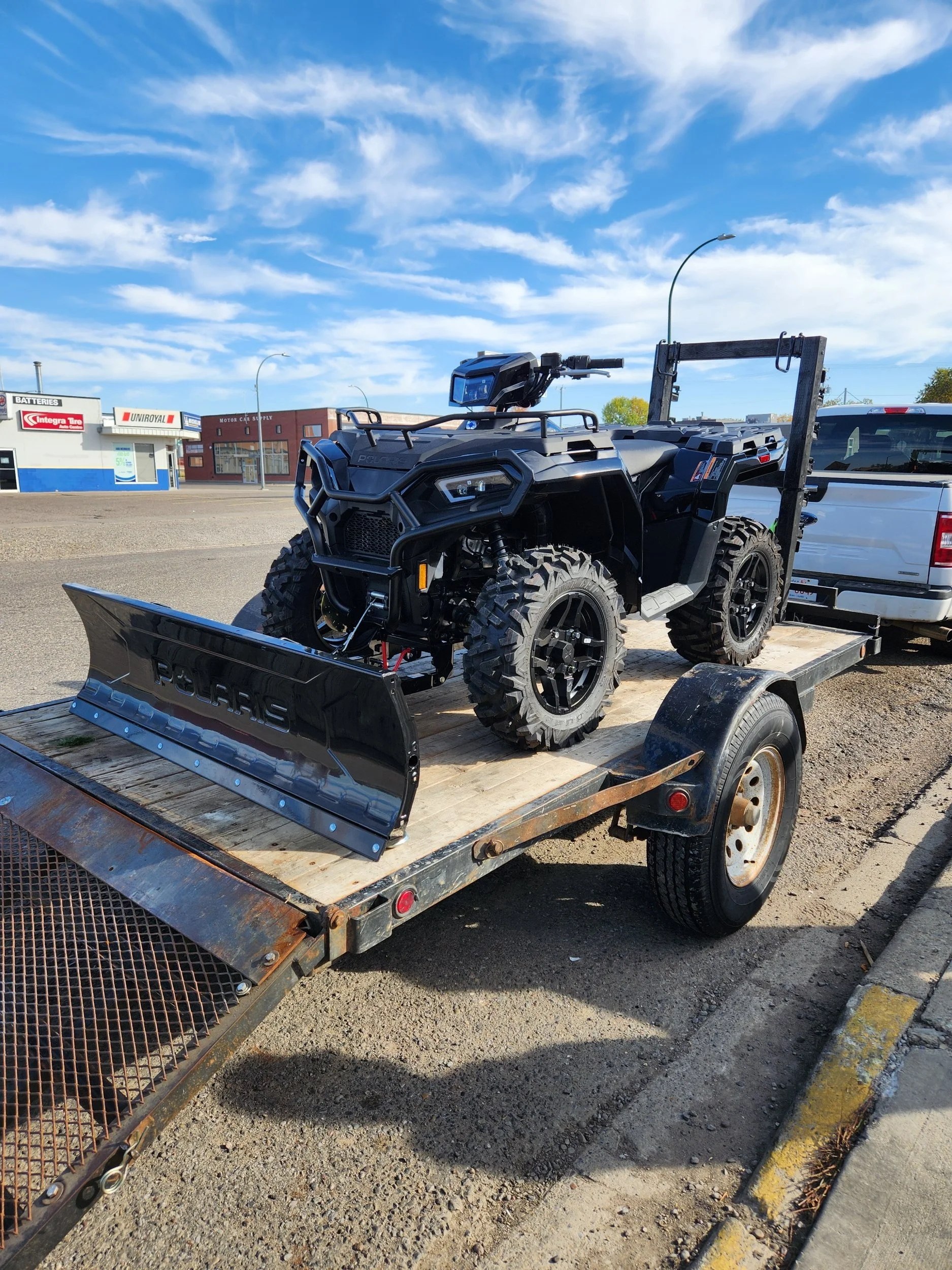 A black Polaris off-road vehicle with a snow plow attachment on a flatbed trailer, parked on a street with commercial buildings in the background.