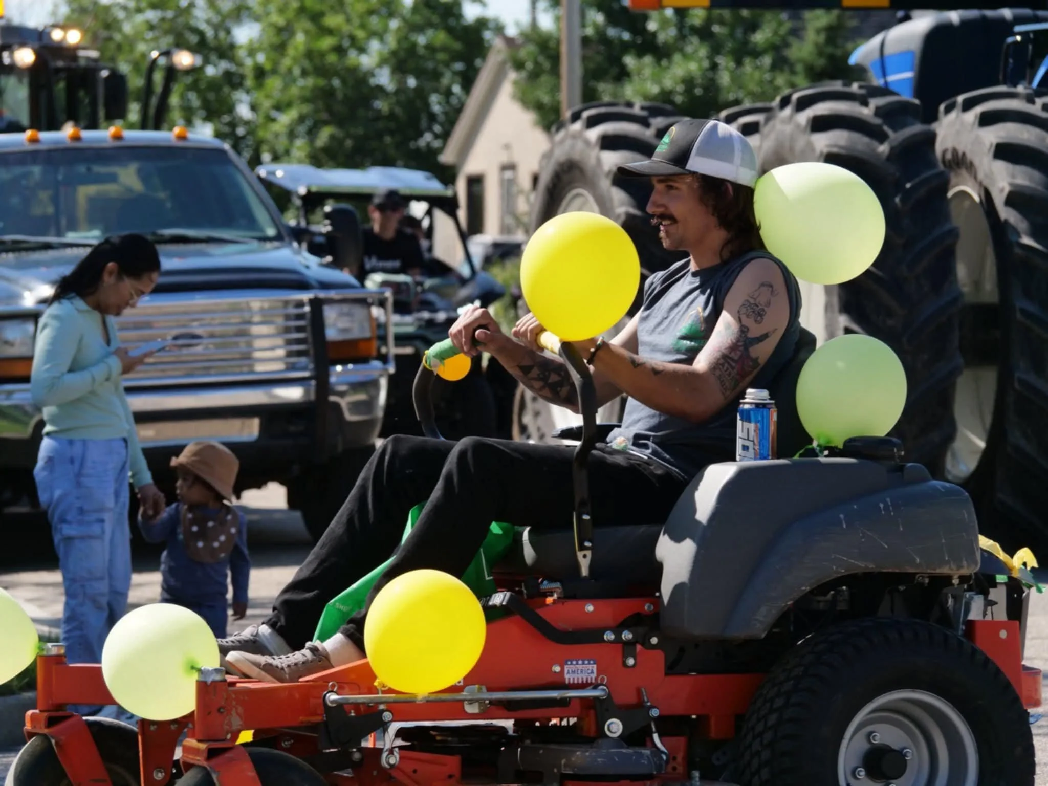 A man with tattoos on his arms sitting on a red lawn mower decorated with yellow and green balloons, wearing a gray sleeveless shirt and a baseball cap. A woman and a small child are in the background, with the woman looking at her phone and the child holding her hand. Several large tractor tires and vehicles are visible in the background.