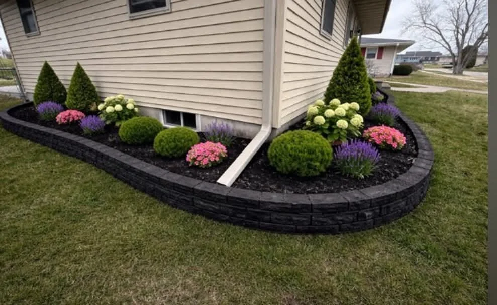 landscaped flower bed with shrubbery and flowers around the base of a beige house with a curved black brick border
