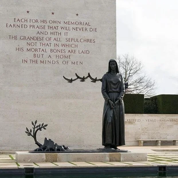 A memorial featuring a statue of a woman in a long dress, standing on a stone platform with a tree and greenery behind her. To her left, there is a sculpted tree and rock. Behind the statue, an inscribed wall displays a poem about remembrance and tribute, with a small flock of bird sculptures flying across the wall.