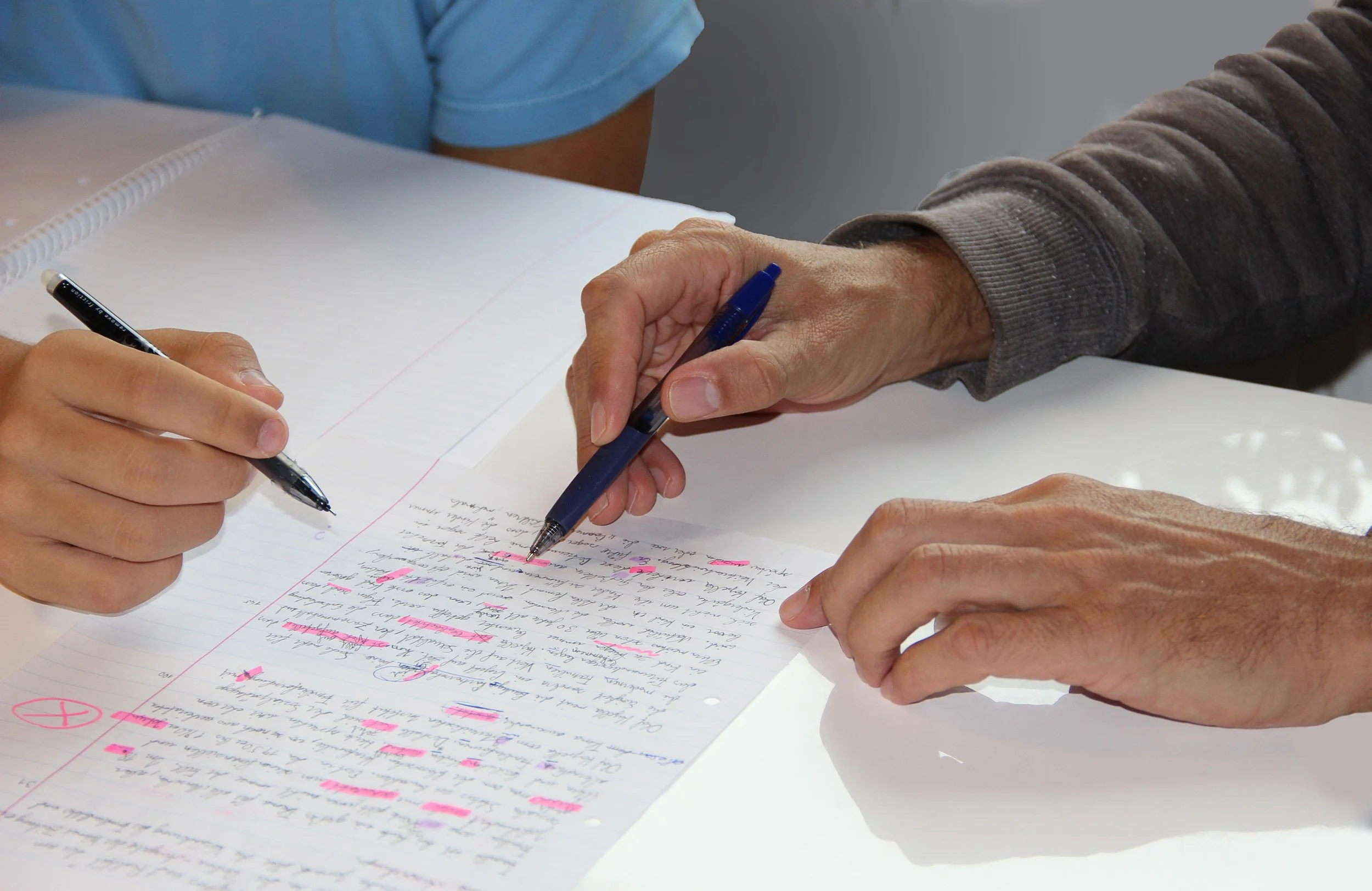 Two people reviewing handwritten notes on a sheet of paper with red marker highlights, one person holding a black pen and the other pointing with a blue pen.