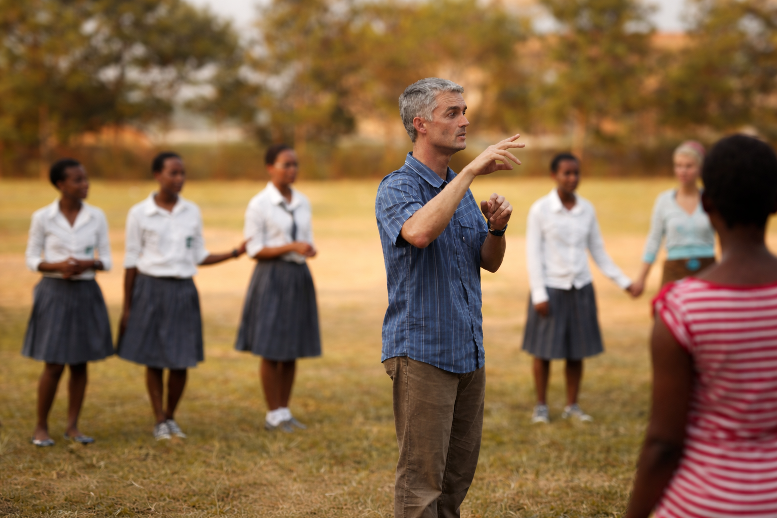 A man speaking to a group of young women outdoors on a grassy field with trees in the background.