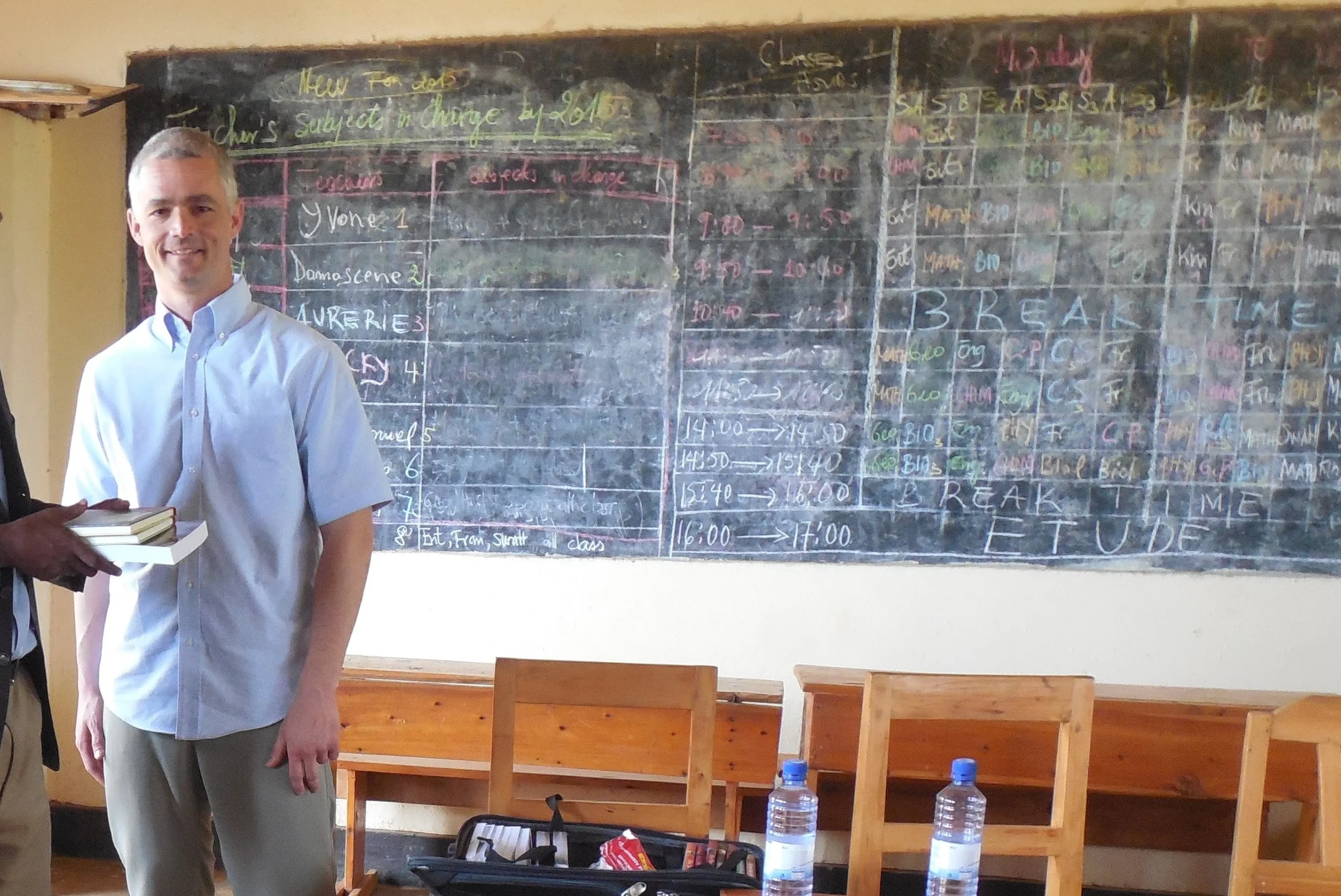 A man standing in front of a classroom blackboard, smiling, with a person next to him holding a stack of books. The blackboard has writing and schedules in various colors, indicating a classroom or school setting. There are bottles of water and chairs in the foreground.