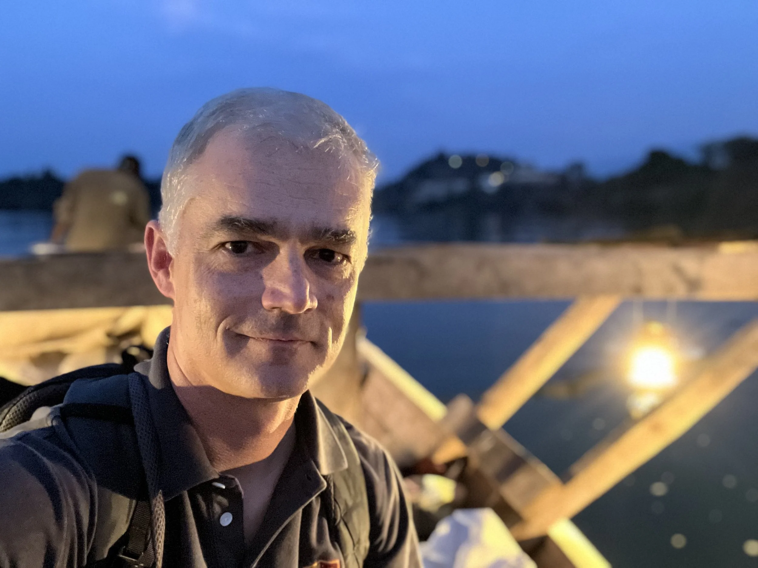 A man with gray hair taking a selfie on a bridge during the evening near a body of water.