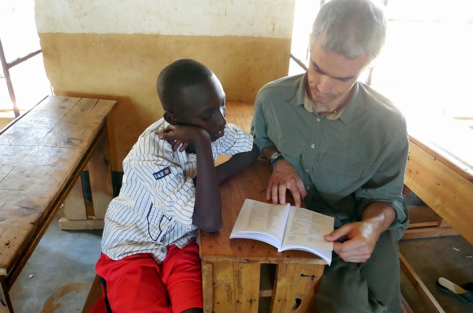 A young boy and an older man sitting at a wooden desk, looking at an open book together in a classroom with wooden benches and a yellow wall.