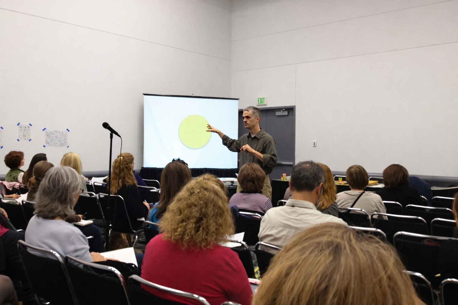 A man in a gray shirt is giving a presentation to an audience of adults seated in black chairs. He is pointing at a large screen with a yellow circle on it. The room has plain white walls and an exit door in the background.