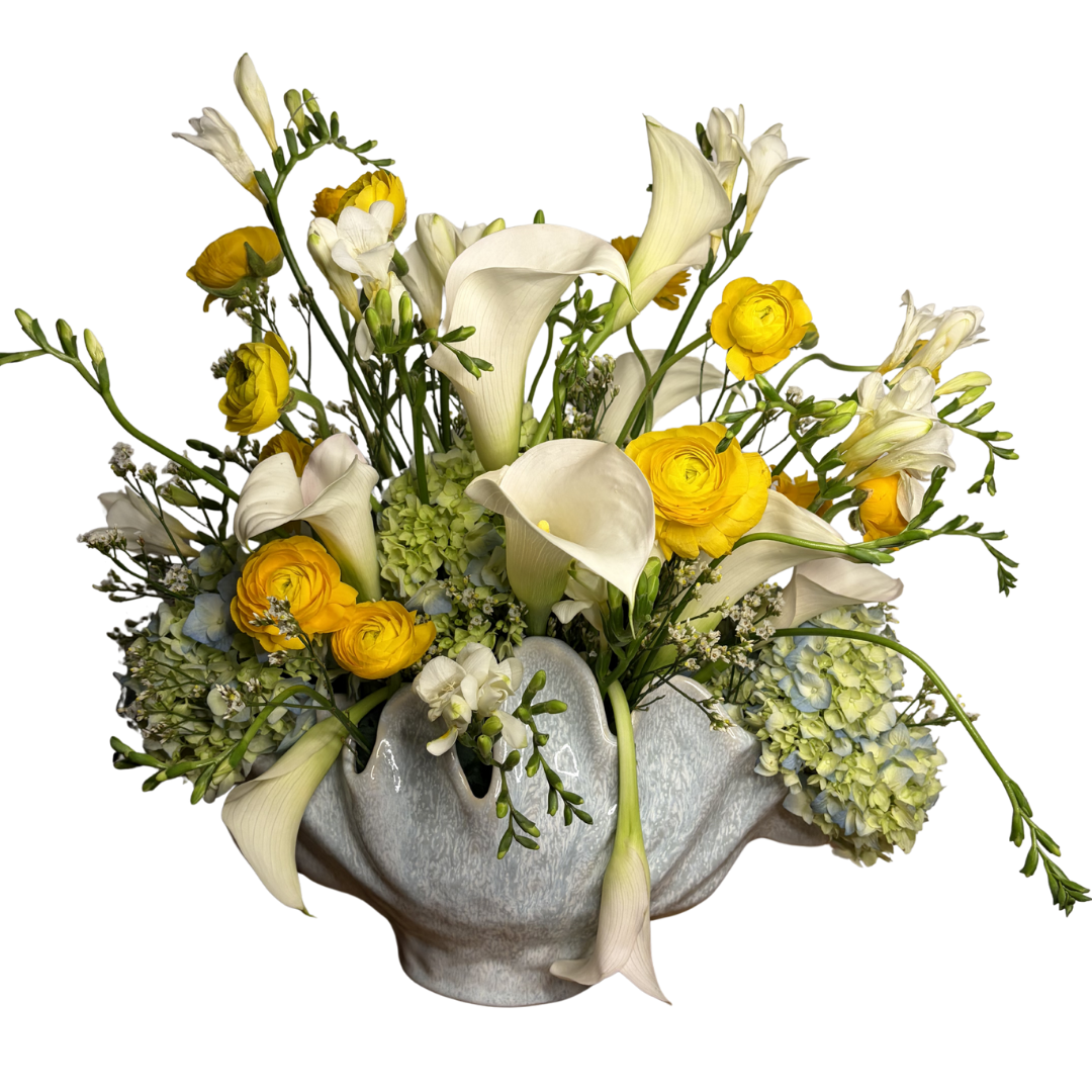 A floral arrangement featuring white calla lilies, yellow ranunculus, white daisies, and other white and green flowers in a white ceramic vase.