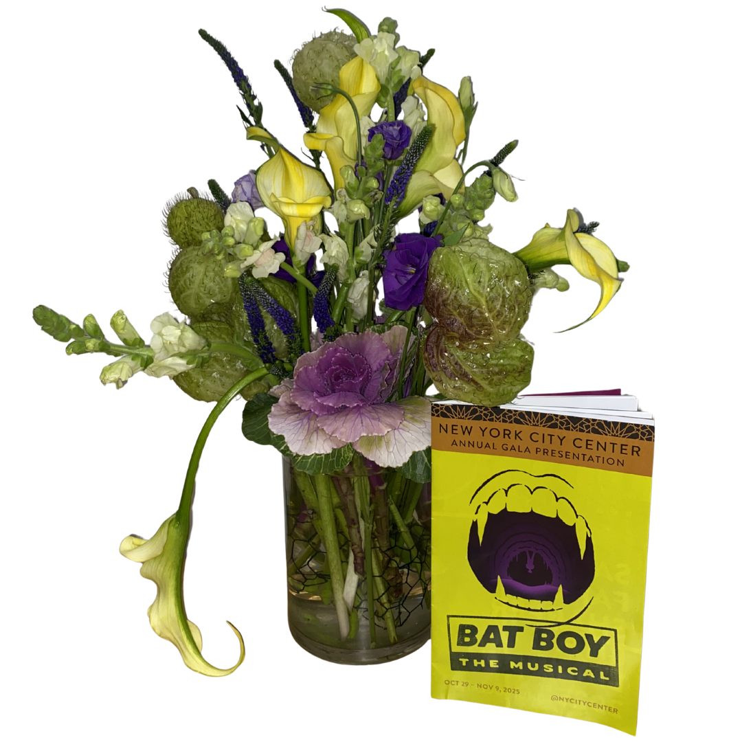 A colorful flower bouquet with calla lilies, violets, and other flowers in a glass vase, accompanied by a yellow promotional leaflet for 'Bat Boy the Musical' at the New York City Center.