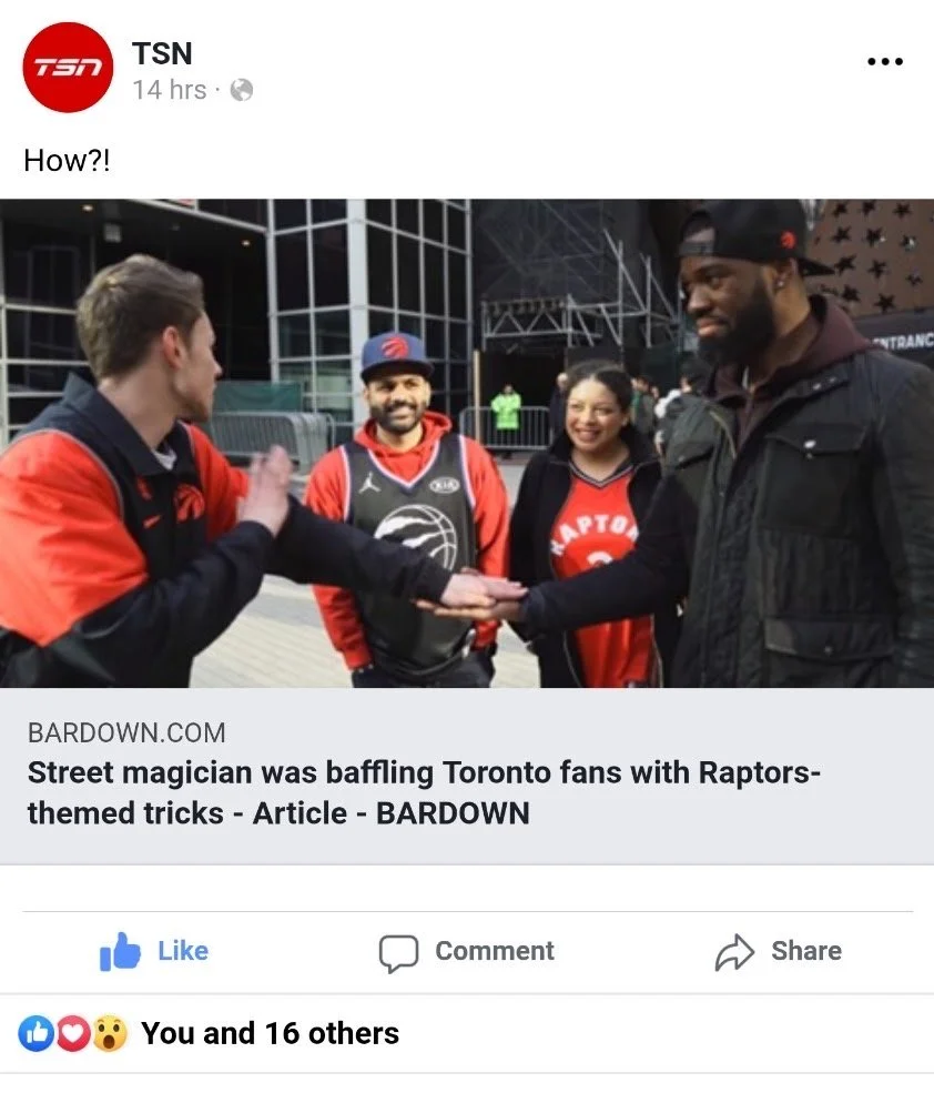 A street magician performing a trick for Toronto Raptors fans. The magician is holding out his hand to another man, who is also extending his hand. Two other fans, a man and a woman, are standing behind them, smiling. All are dressed in Toronto Raptors apparel.