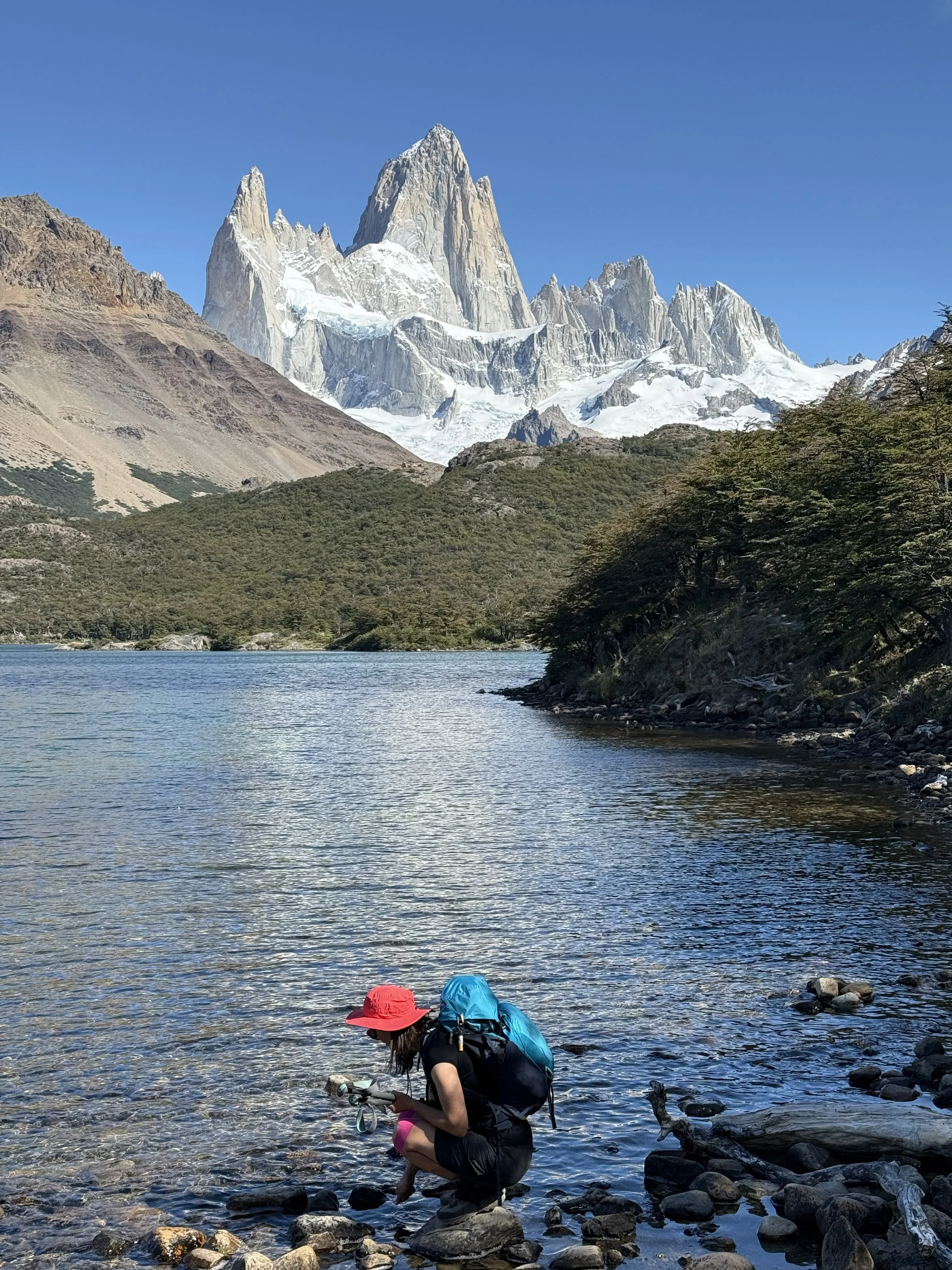 An explorer with a red hat and backpack crouching by a rocky lakeshore, picking up rocks, with mountains and a snow-capped peak in the background.