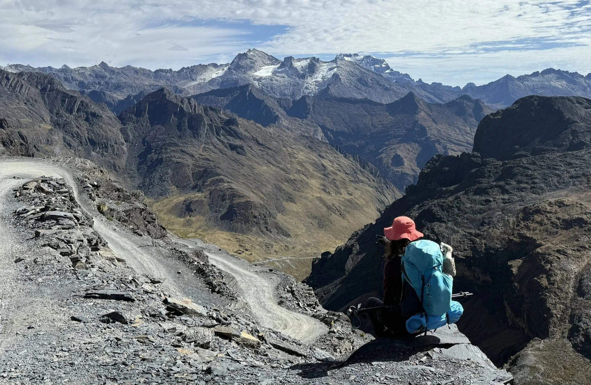 A contemplative woman with a pink hat and a blue backpack sitting on a rocky ledge enjoying the view, in a mountainous landscape with rugged peaks and a winding trail.