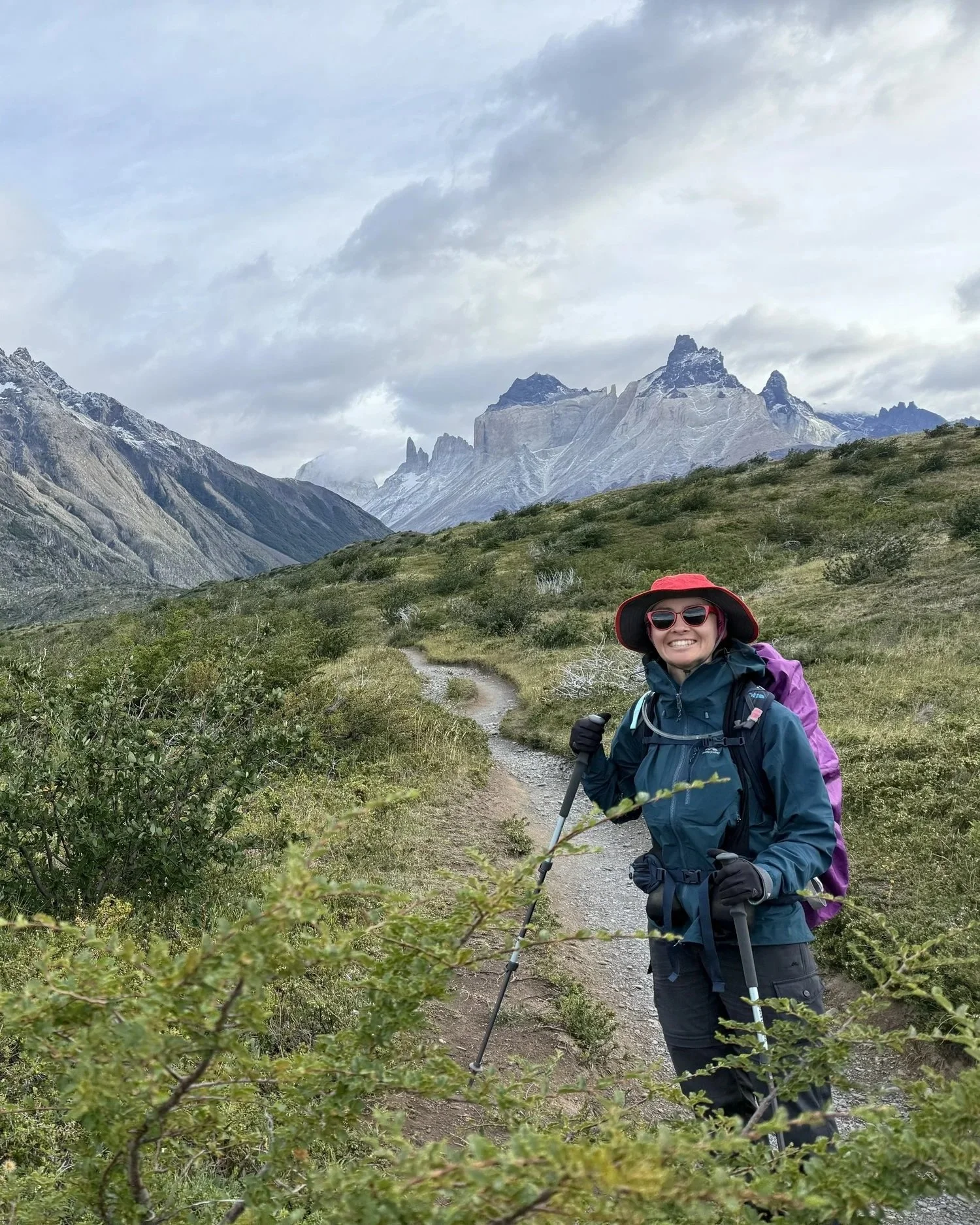 A smiling woman with hiking gear, including a purple backpack, red hat, sunglasses, and hiking poles, standing on a trail in a mountainous landscape with green shrubs and rocky peaks spreading out in the distance.