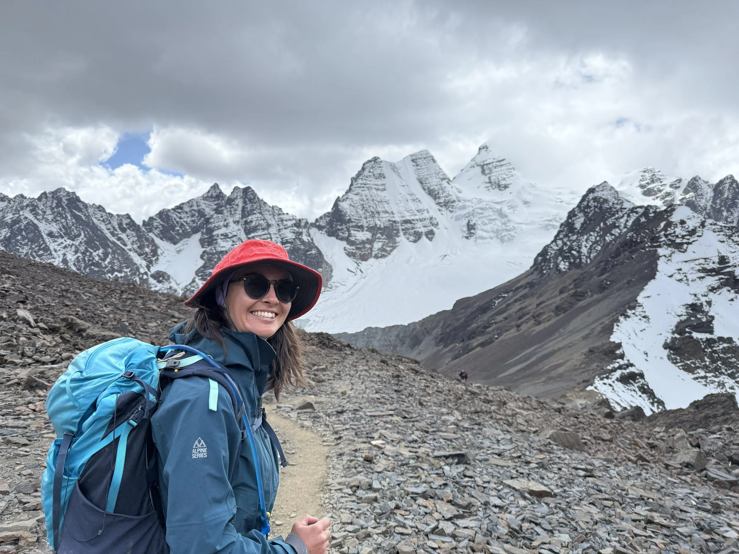 A woman going on an adventure, in outdoor hiking gear, including sunglasses, a red hat, and a blue jacket, smiling while standing on a rocky mountain trail with snow-covered peaks in the background.