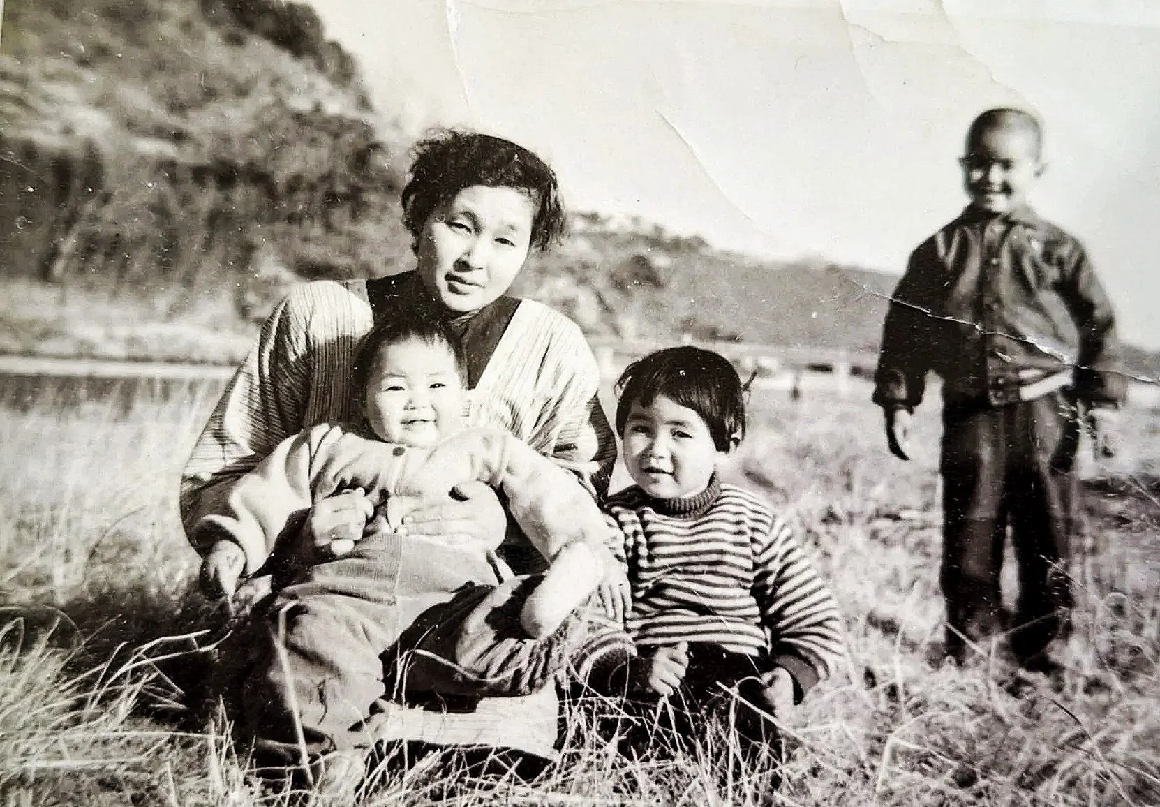 An old black and white photo of a Japanese mother of three children outdoors in a field with hills in the background. The woman is sitting and holding a young baby, while a toddler sits next to her. Her son stands a few feet away in the background.