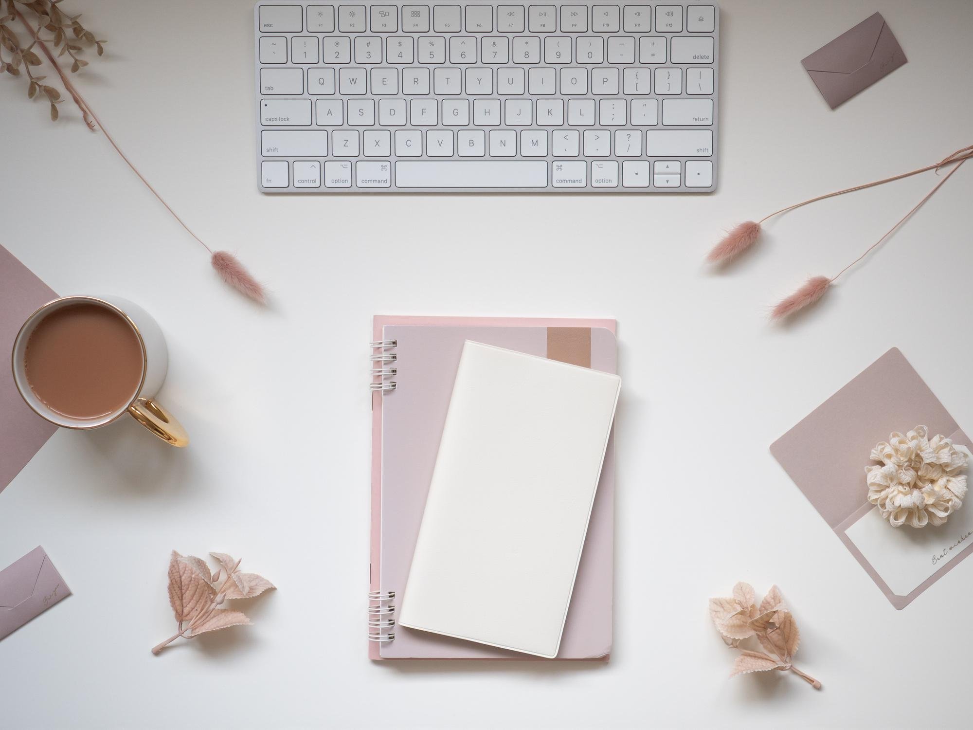 A white desk with a white keyboard, a cup of coffee, pink and beige decorative plants, a pink notebook, and a small envelope.