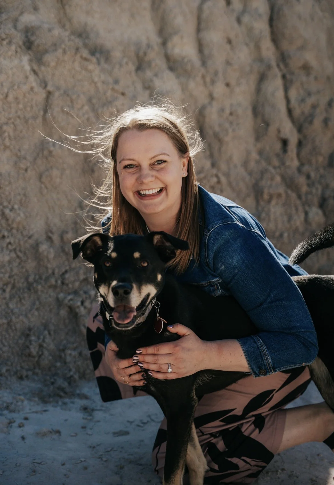 A young woman smiling and hugging a black and tan dog on a rocky outdoor surface.