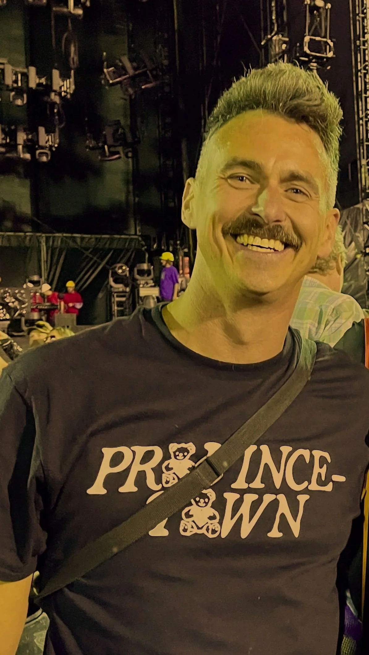 Smiling man with a beard and mustache wearing a black T-shirt with paw print graphics and the words 'Prince Town' on it. Background shows a stage with black backdrop, lighting equipment, and two people in hard hats.
