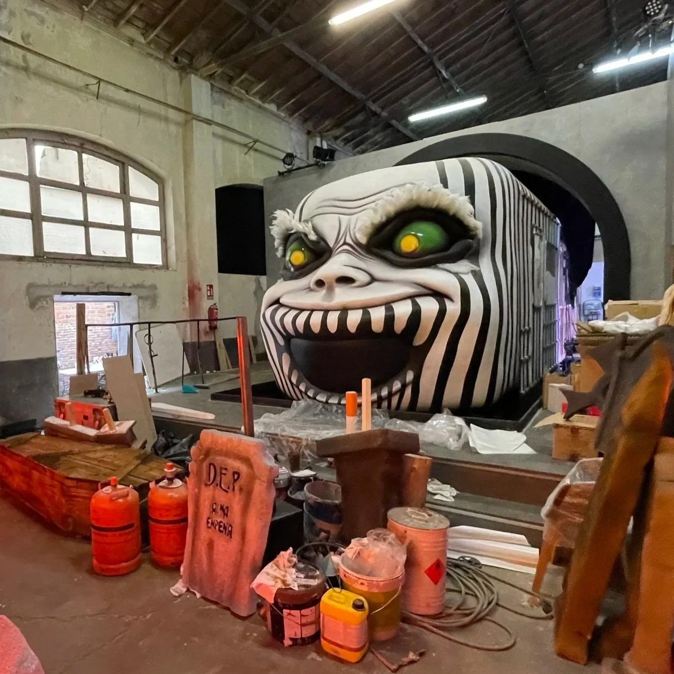 A large, cartoonish face with a wide-open mouth, big yellow eyes, and black and white striped patterns covers a structure in a workshop or warehouse setting. Construction materials and tools are scattered in the foreground.