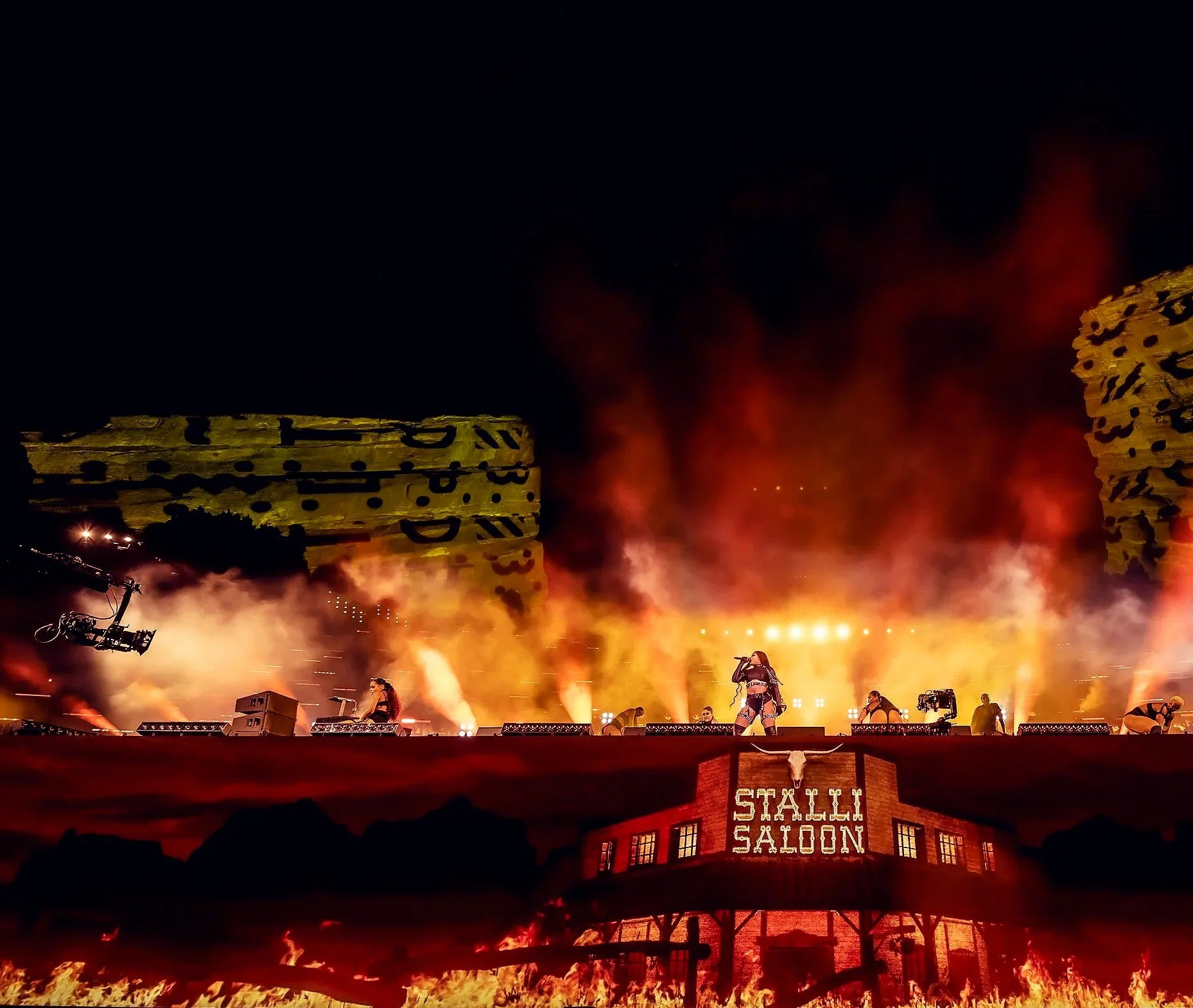A stage performance at a Western-themed Saloon with large flames and smoke, a performer in center with microphone, and a sign reading 'STALUM SALOON' on a building facade, in front of a desert landscape with mountains at night.