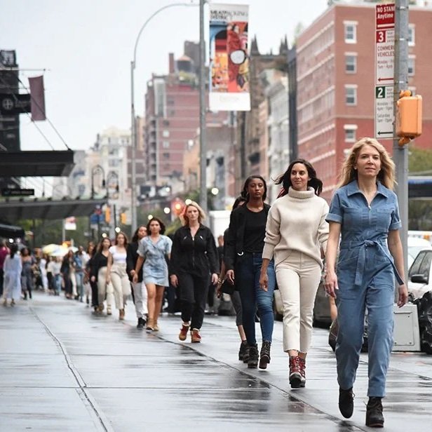 A line of women walking on a city sidewalk on a rainy day with buildings and signs in the background.