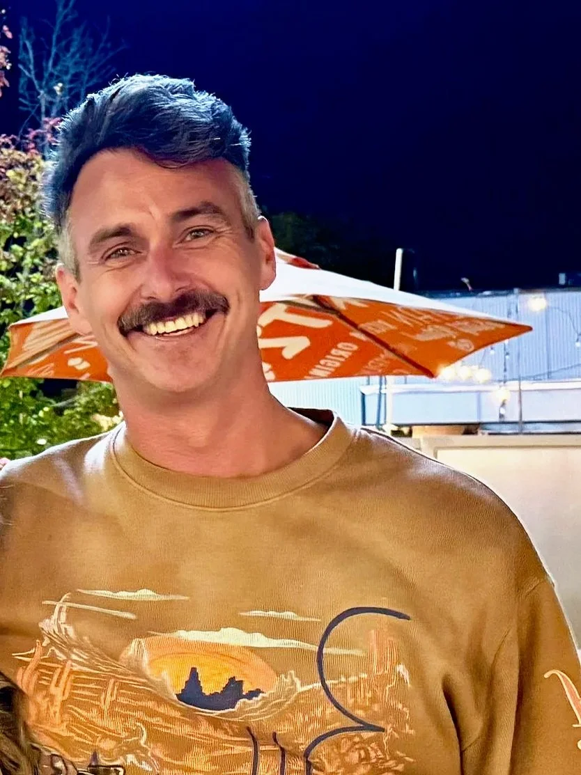 Smiling man with a mustache and styled hair, wearing a tan T-shirt with a sunset graphic, outdoors at night with umbrella and trees in background.