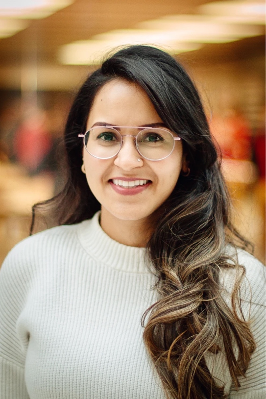 A woman with long, dark hair and glasses smiling indoors with blurred warm-colored background.