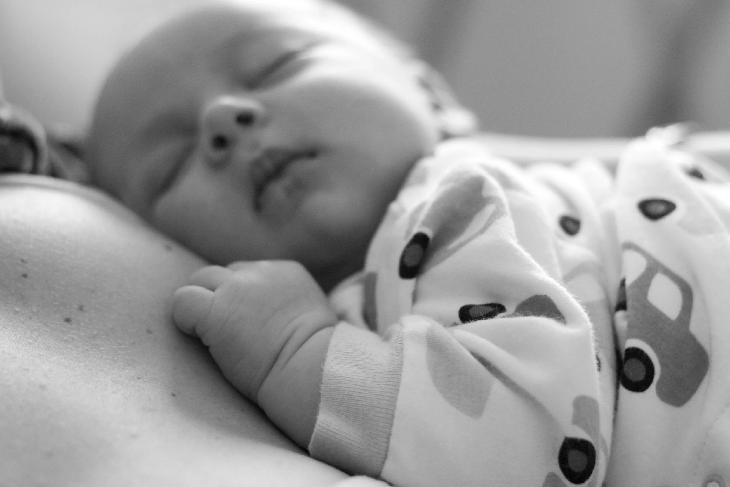 A sleeping baby lying on a person's chest, gripping the person's finger, in a hospital setting, in black and white.
