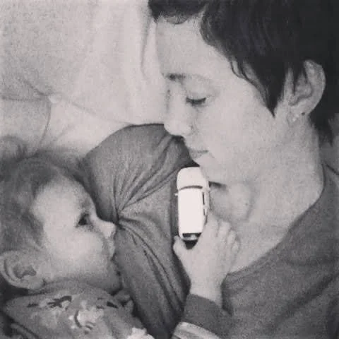 A young woman and a baby lying close together, with the woman gently holding the baby's hand and the baby touching her face, both appearing to look at each other.