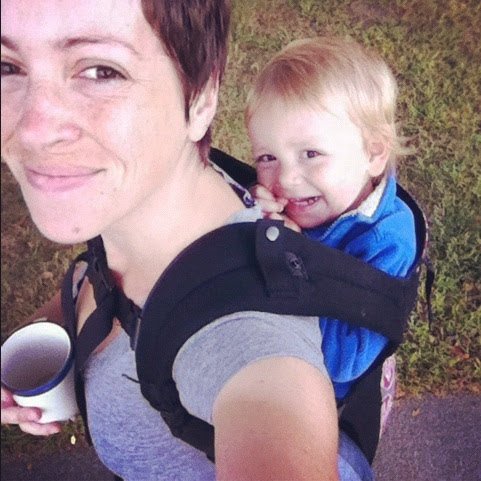 A young woman smiling while carrying a small child in a backpack carrier on her back, outdoors on a grassy area.