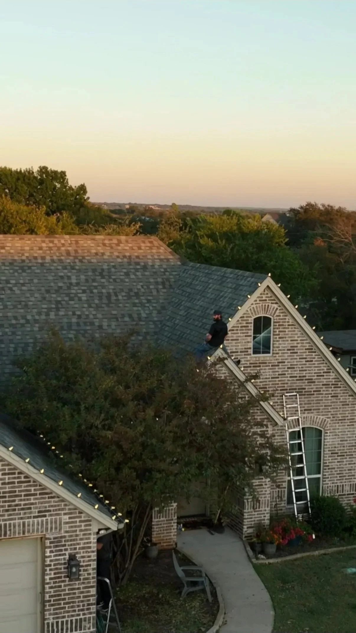 Aerial view of Bearded Elf Co. owner Weston Rutkowski installing Christmas lights on brick home rooftop in Granbury Texas — professional holiday light installation ladder on roof DFW