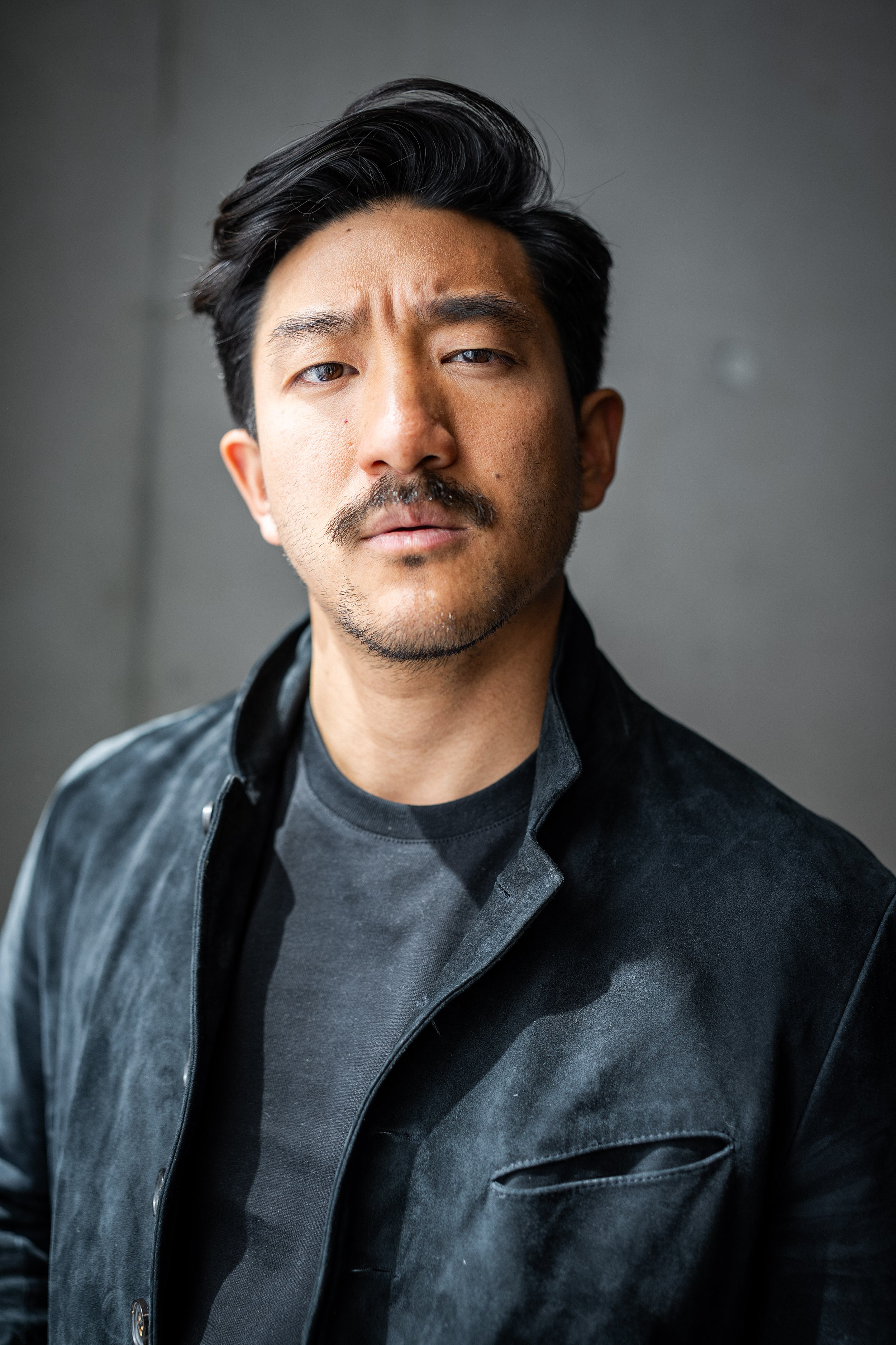Close-up portrait of a young Asian man with short black hair and light facial hair, wearing a dark blue denim shirt, against a gray background.
