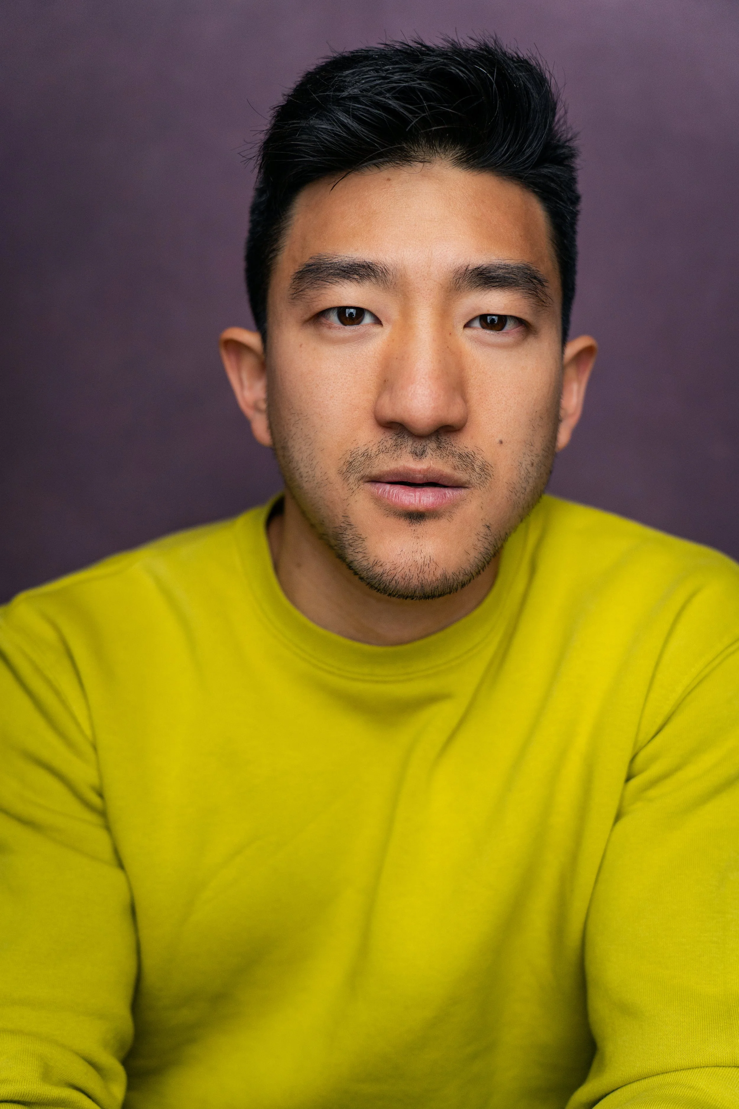 A young man with black hair, wearing a yellow shirt, looking directly at the camera with a neutral expression, against a dark purple background.
