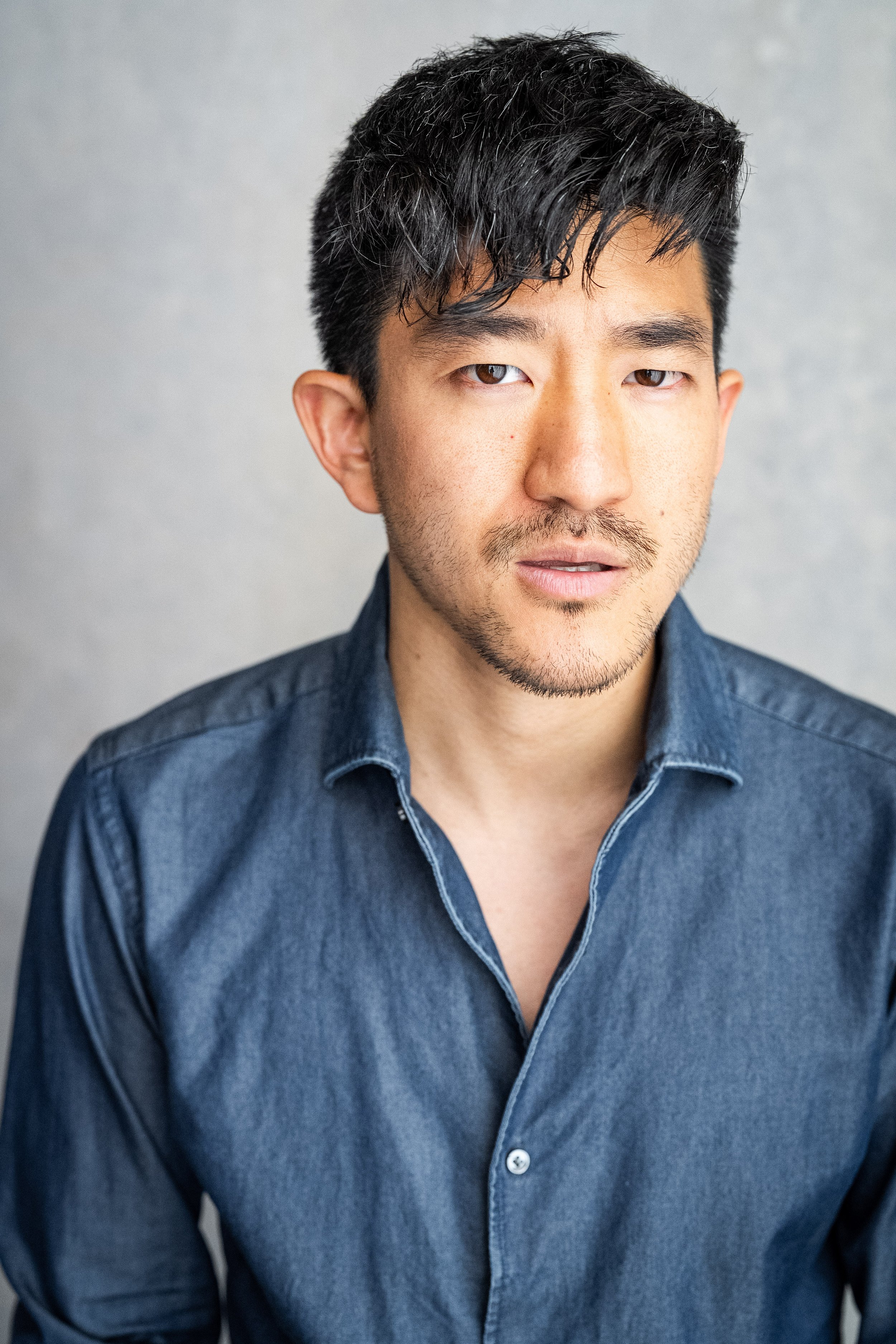 Close-up portrait of a young Asian man with short black hair and light facial hair, wearing a dark blue denim shirt, against a gray background.