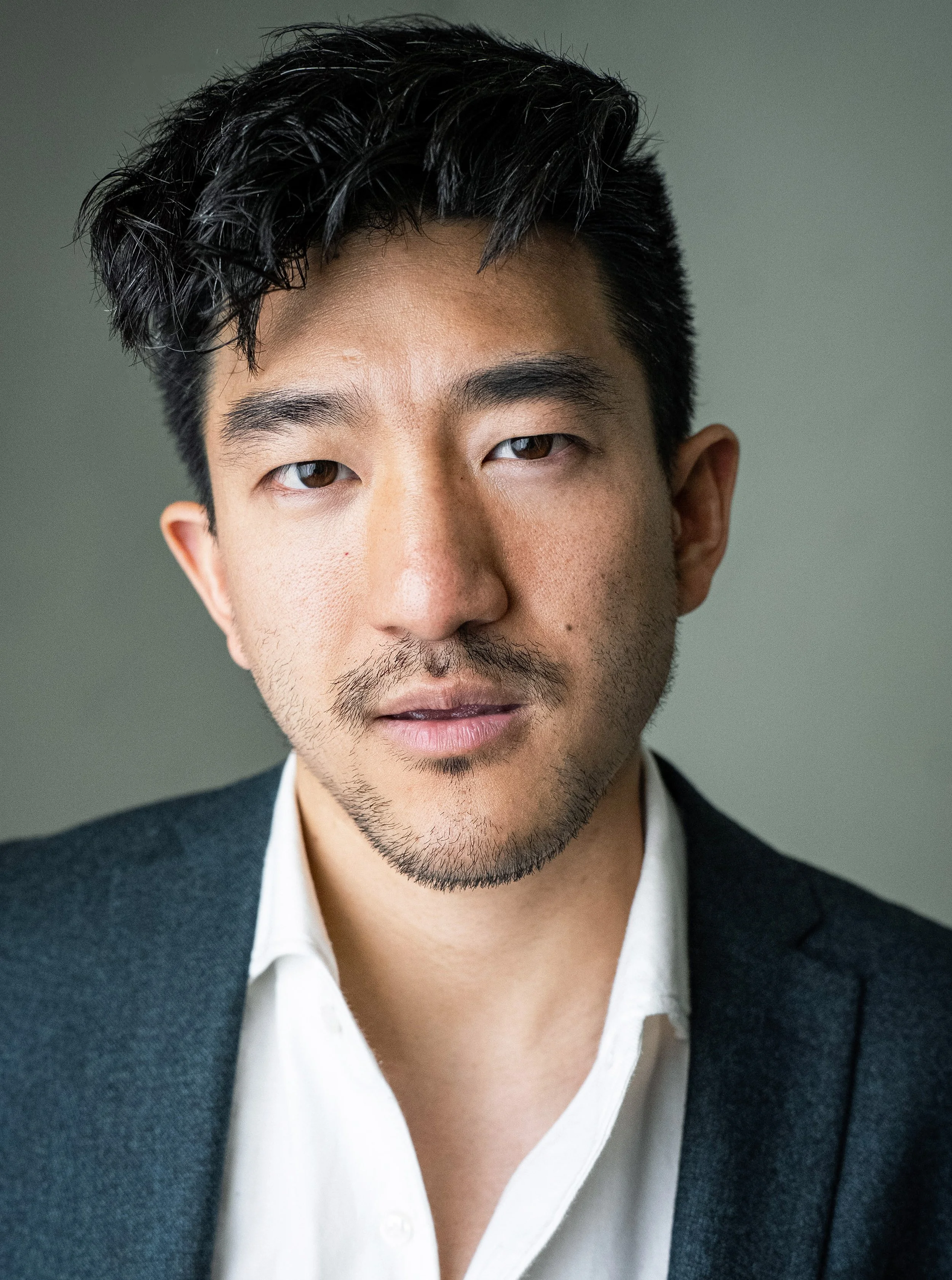 Portrait of a young Asian man with dark, wavy hair, wearing a dark blazer over a white shirt, against a plain background.