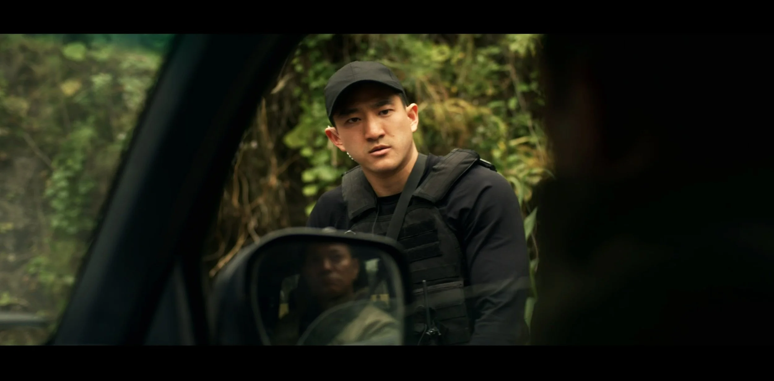 A young man dressed in black with a cap, wearing a tactical vest, is looking at the camera outside a vehicle in a wooded area.