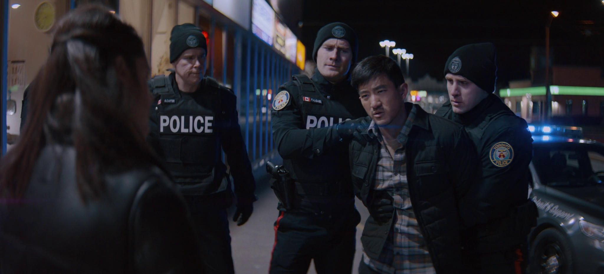 Nighttime scene showing three police officers in tactical gear arresting a man. A woman is in the foreground, facing away from the camera. The officers are focused on the man, and a police car is visible in the background.