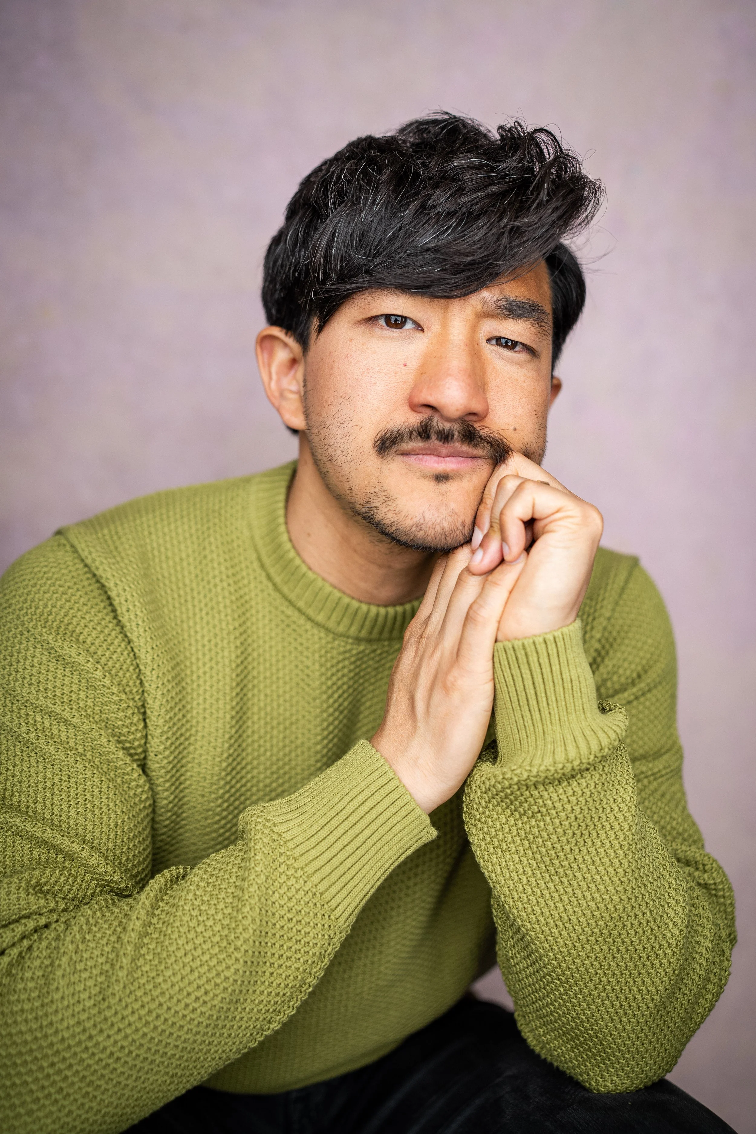 Close-up portrait of a smiling young Asian man with short black hair, wearing a dark shirt over a gray T-shirt, against a plain background.