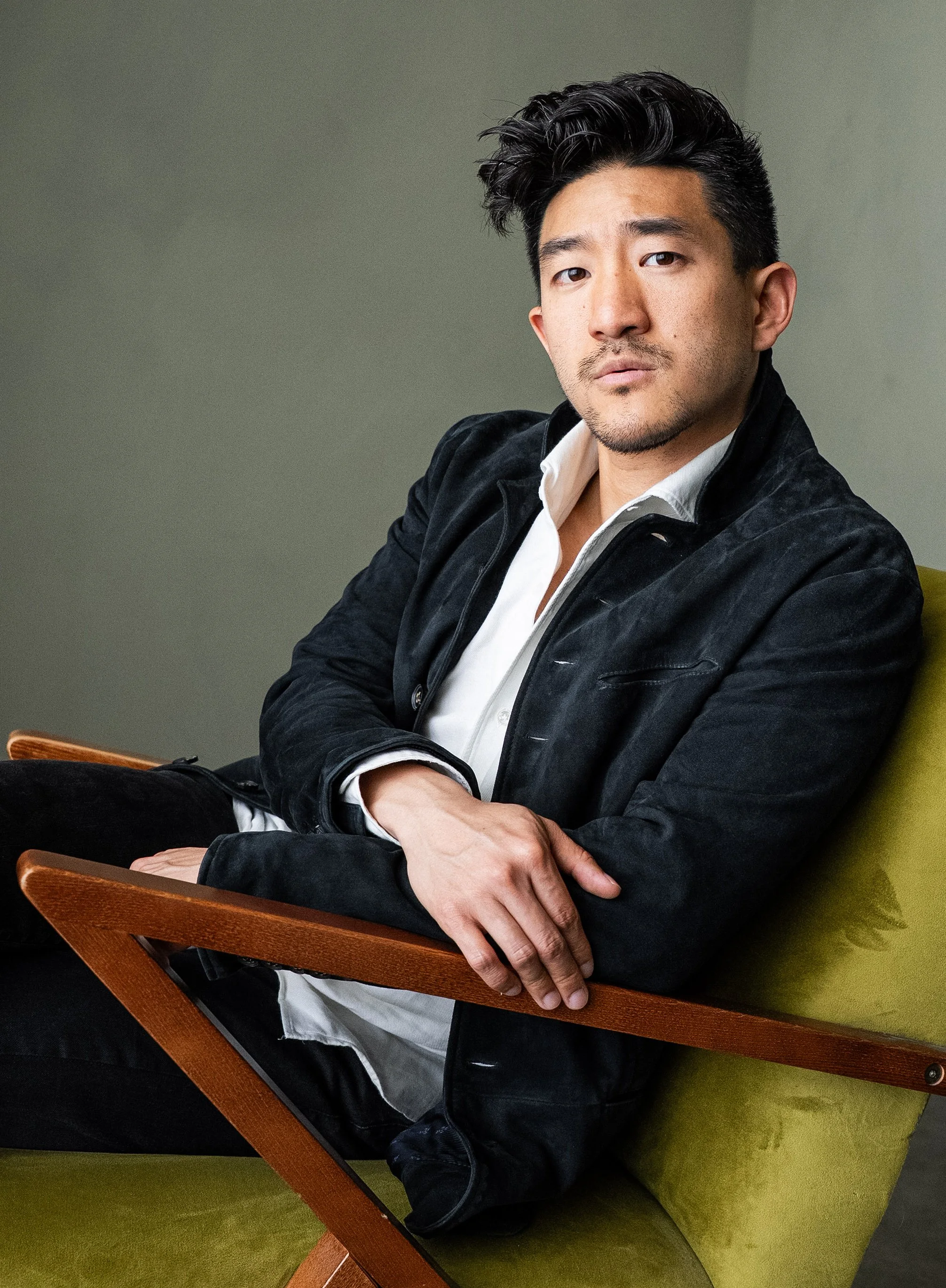 Portrait of a confident man sitting on a green chair, wearing a black jacket and white shirt, with a neutral expression.