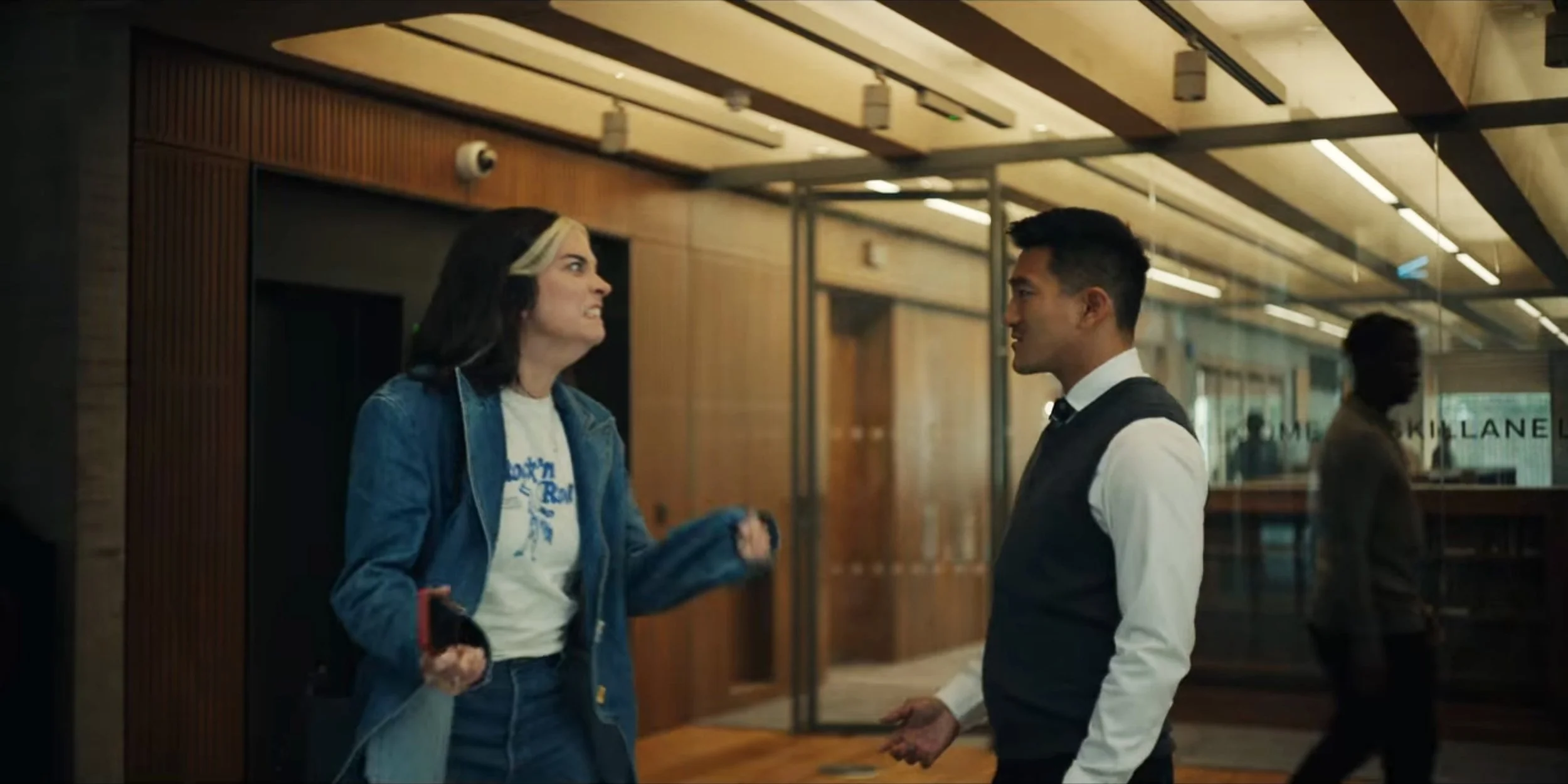 A woman and a man are having an argument inside a modern building, with the woman appearing angry and the man smiling calmly. There are other people in the background walking by.