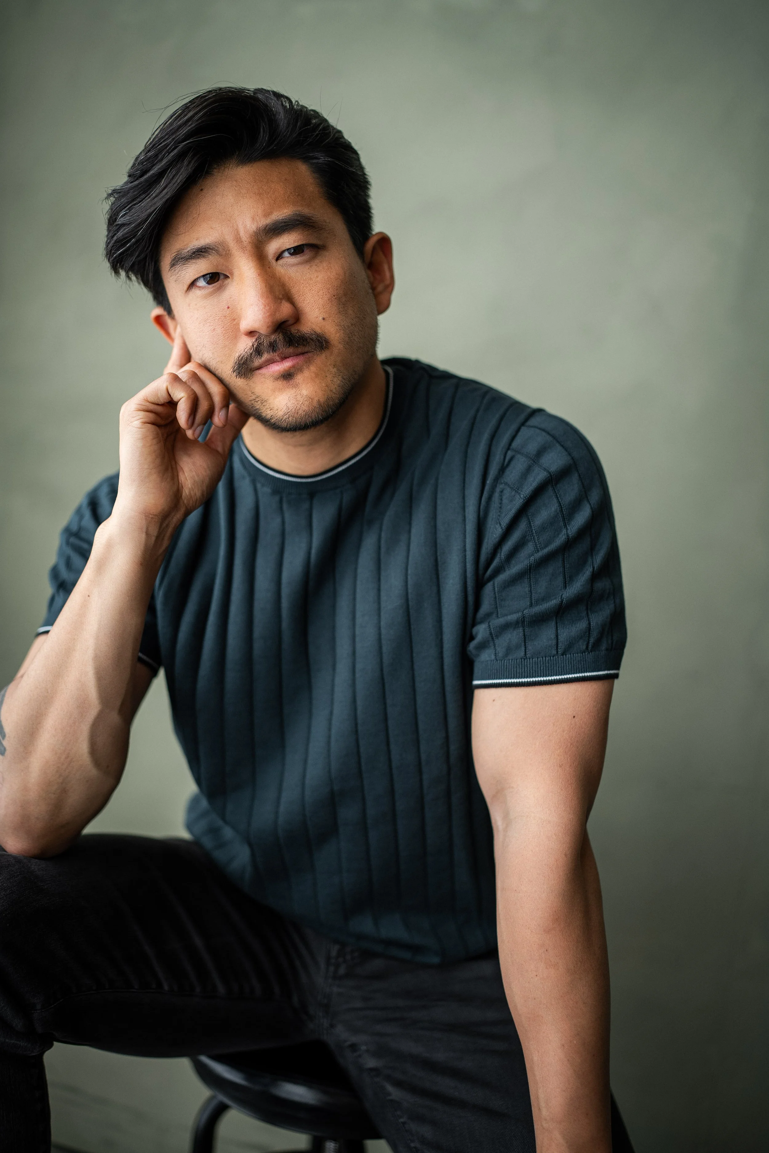 Close-up portrait of a man with black hair, light facial hair, and wearing a brown sweater, against a light green background.