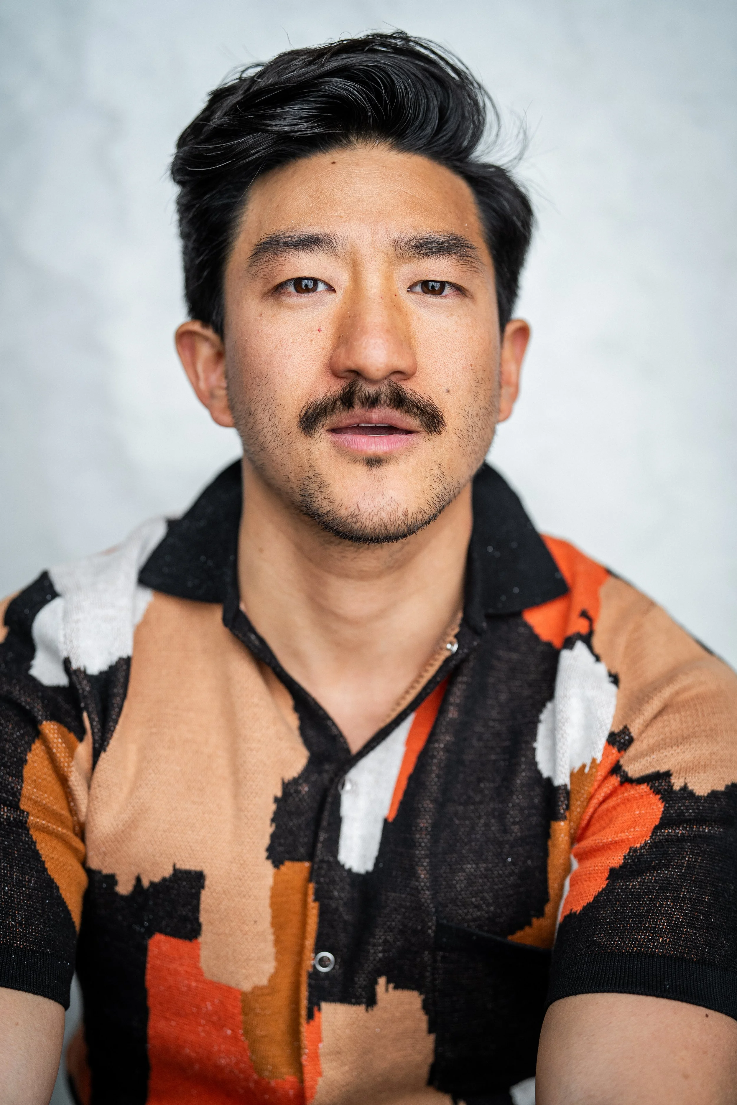 A young man with dark hair, facial hair, and light brown skin, wearing a light green sweater, looking into the camera with a neutral expression, against a plain light background.