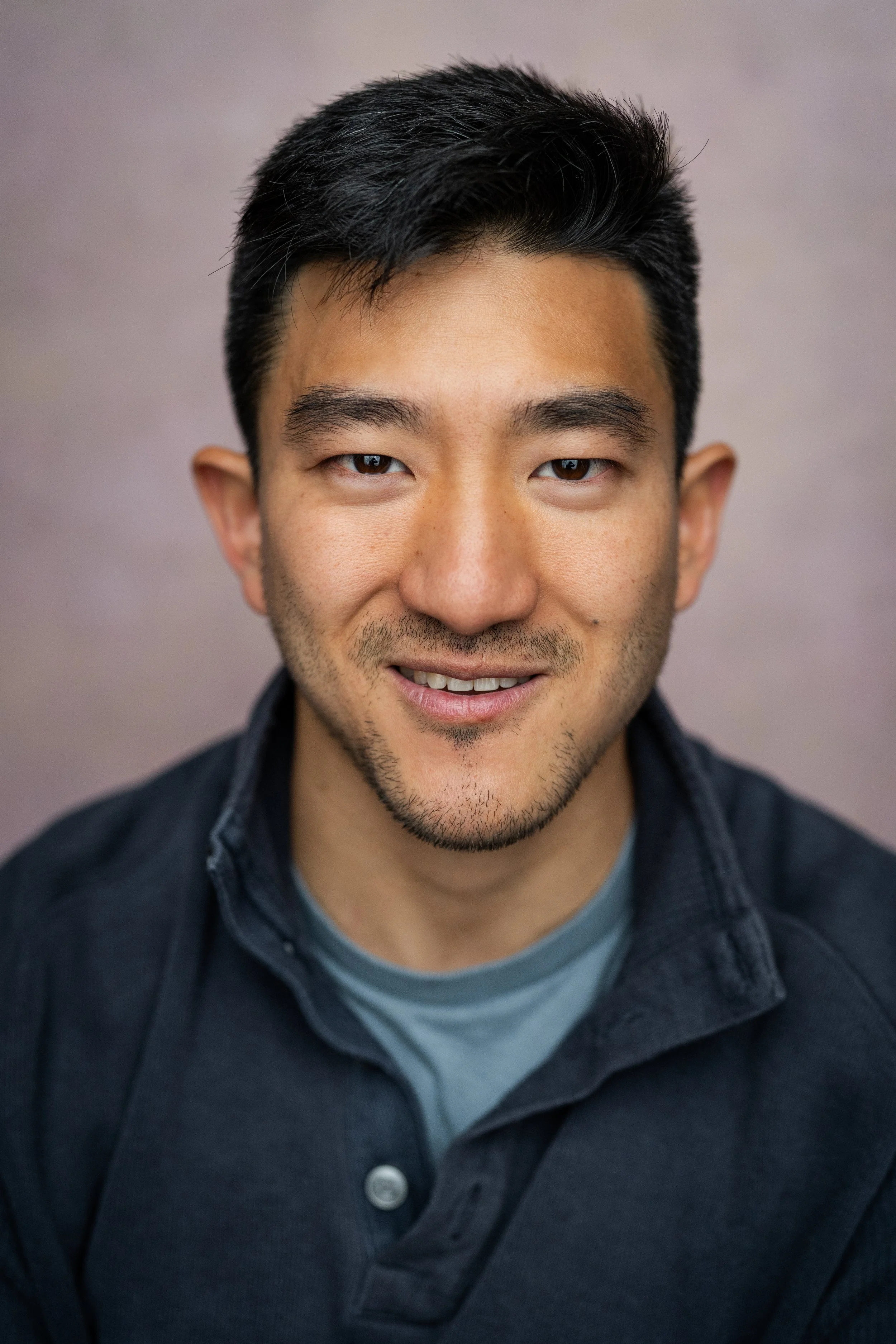 Close-up portrait of a smiling young Asian man with short black hair, wearing a dark shirt over a gray T-shirt, against a plain background.