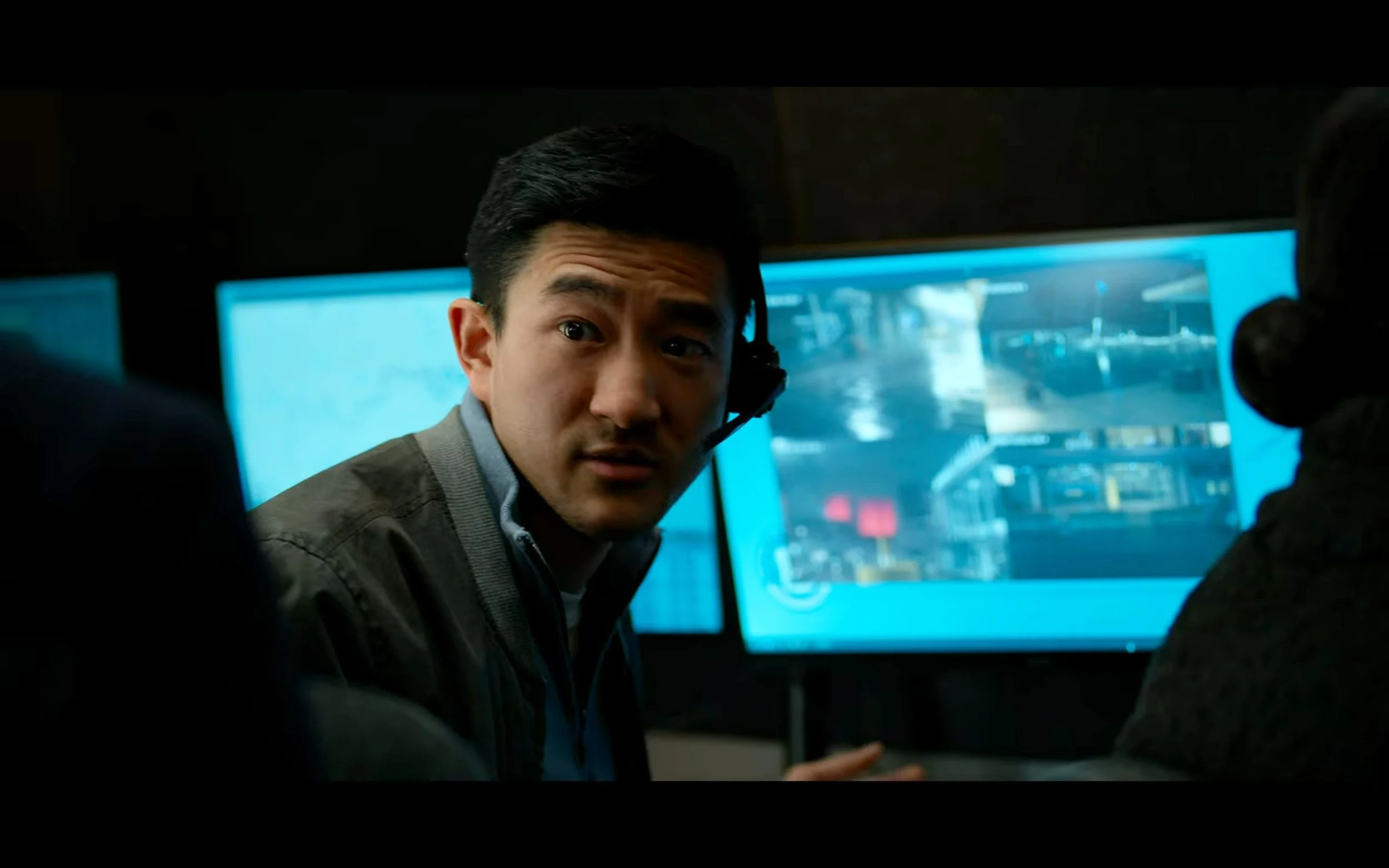 A man wearing a jacket and headset, looking at the camera, in a control room with multiple monitors displaying cityscape scenes.