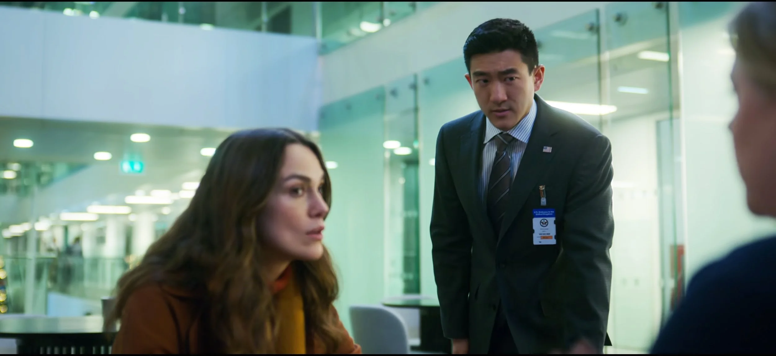 A woman with long brown hair and a man in a suit are seated in an airport, with another man standing between them. The standing man is dressed in a dark suit with an ID badge and appears to be speaking to or confronting the seated woman.