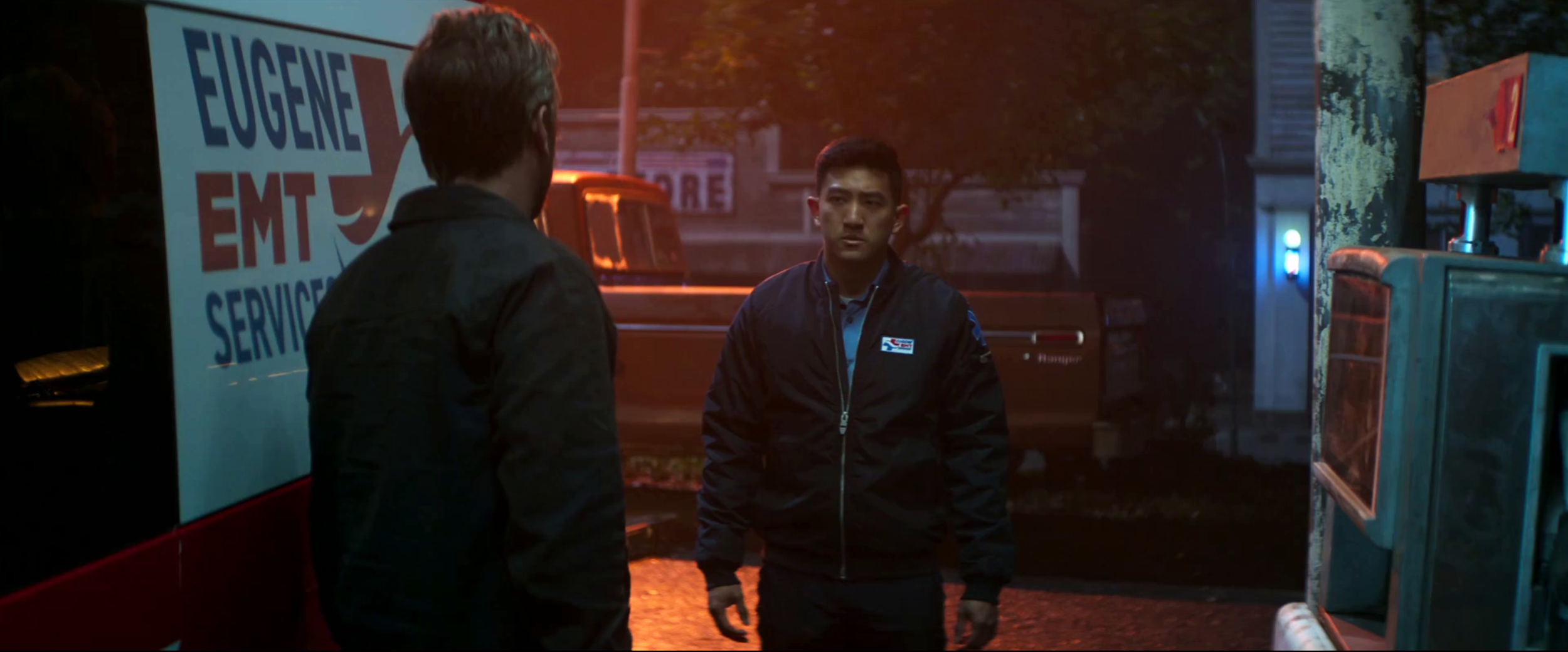 Two men are standing outside on a rainy evening near an emergency medical service vehicle, with one man facing the camera and the other with his back turned, near a public telephone booth.