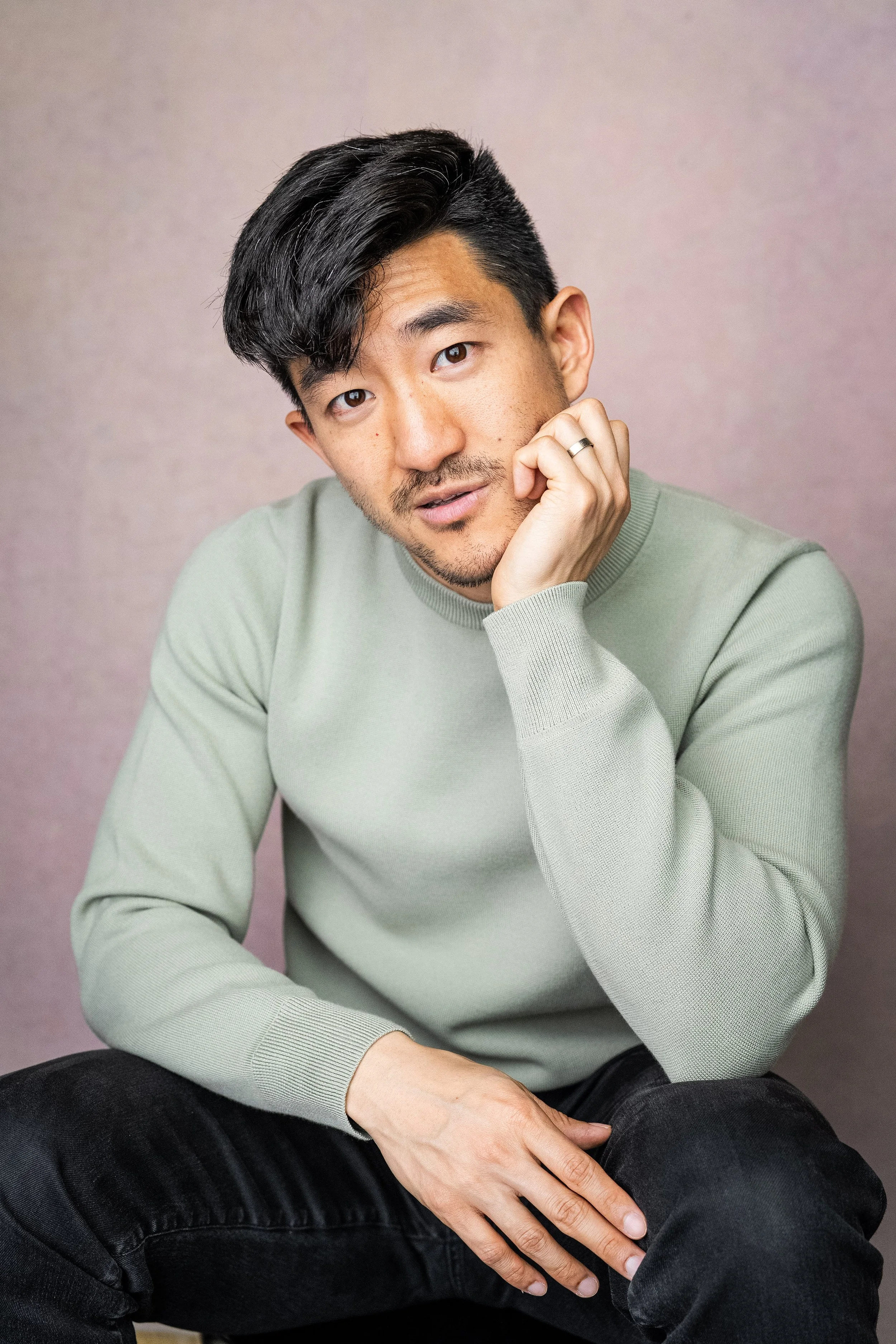 A young Asian man with black hair, slight beard, wearing a light green sweater and black pants, sitting and resting his chin on his hand, looking at the camera with a thoughtful expression against a plain background.